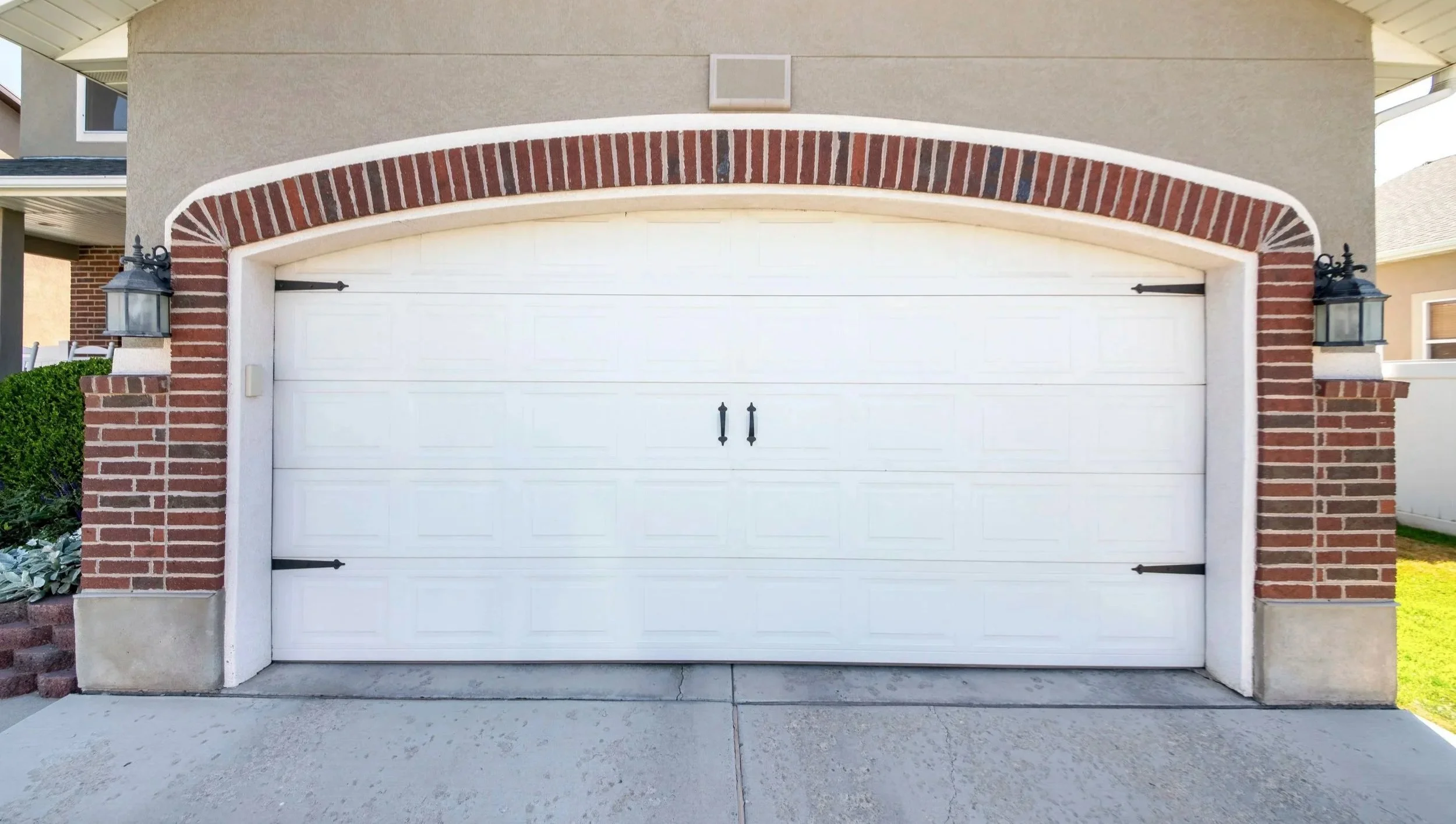 A white garage door with decorative black hinges and handles, framed by brick pillars and a brick archway, in front of a beige stucco house.