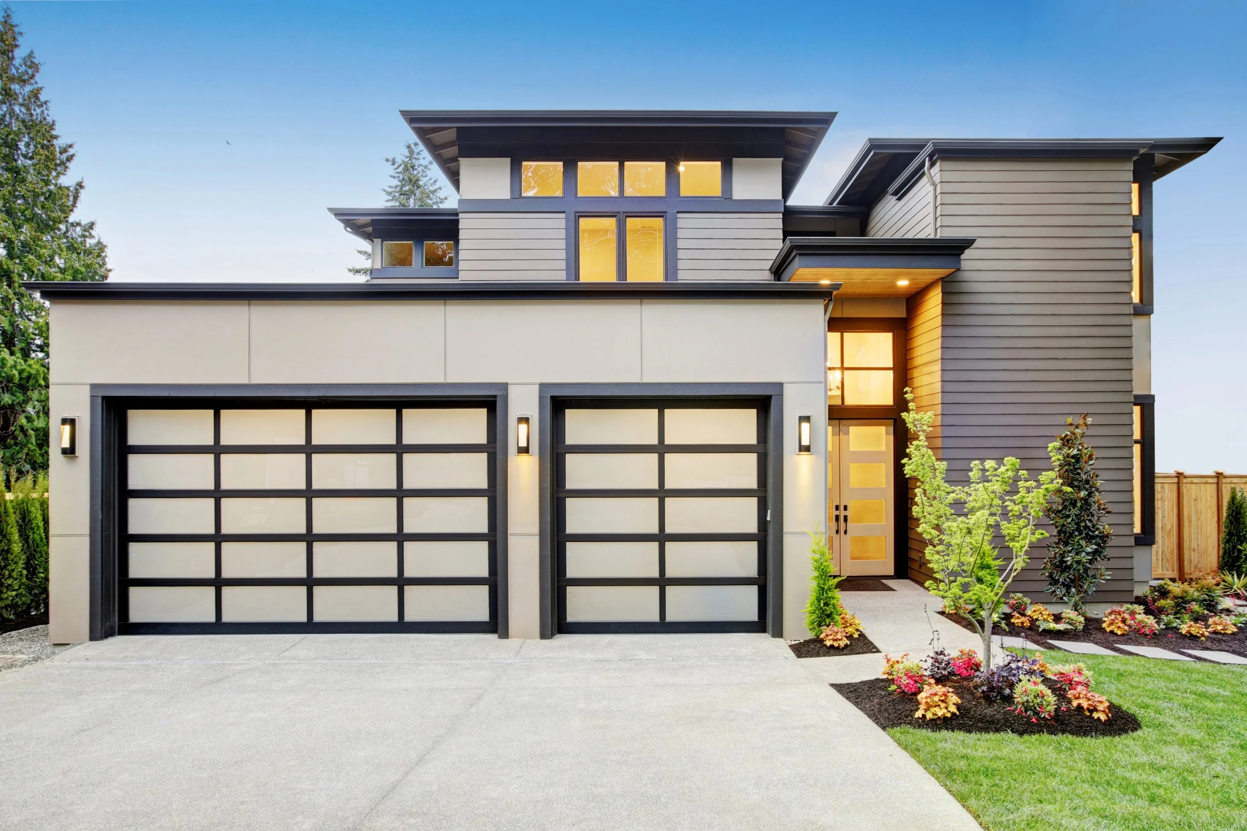 Modern two-story house with gray and white exterior, large windows, two garage doors, a front pathway with landscaped flowers and small trees, and a blue sky background.