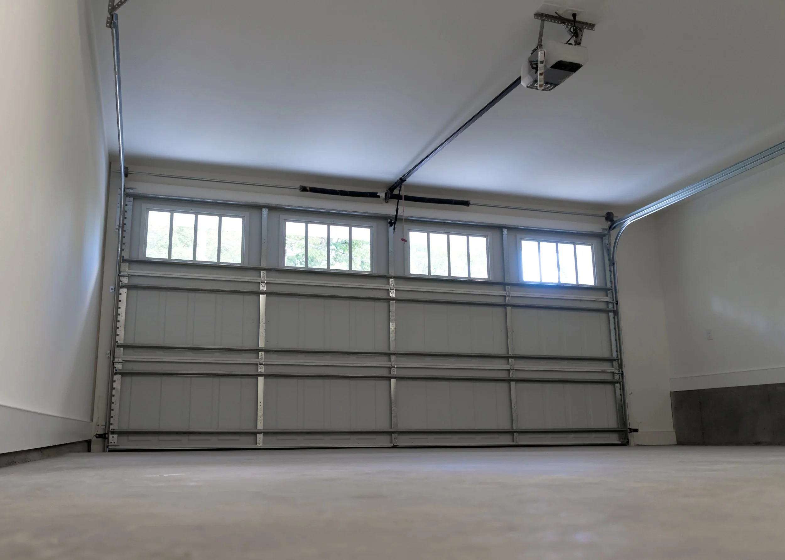 An empty garage with a closed overhead door, windows allowing natural light, and ceiling-mounted garage door opener.