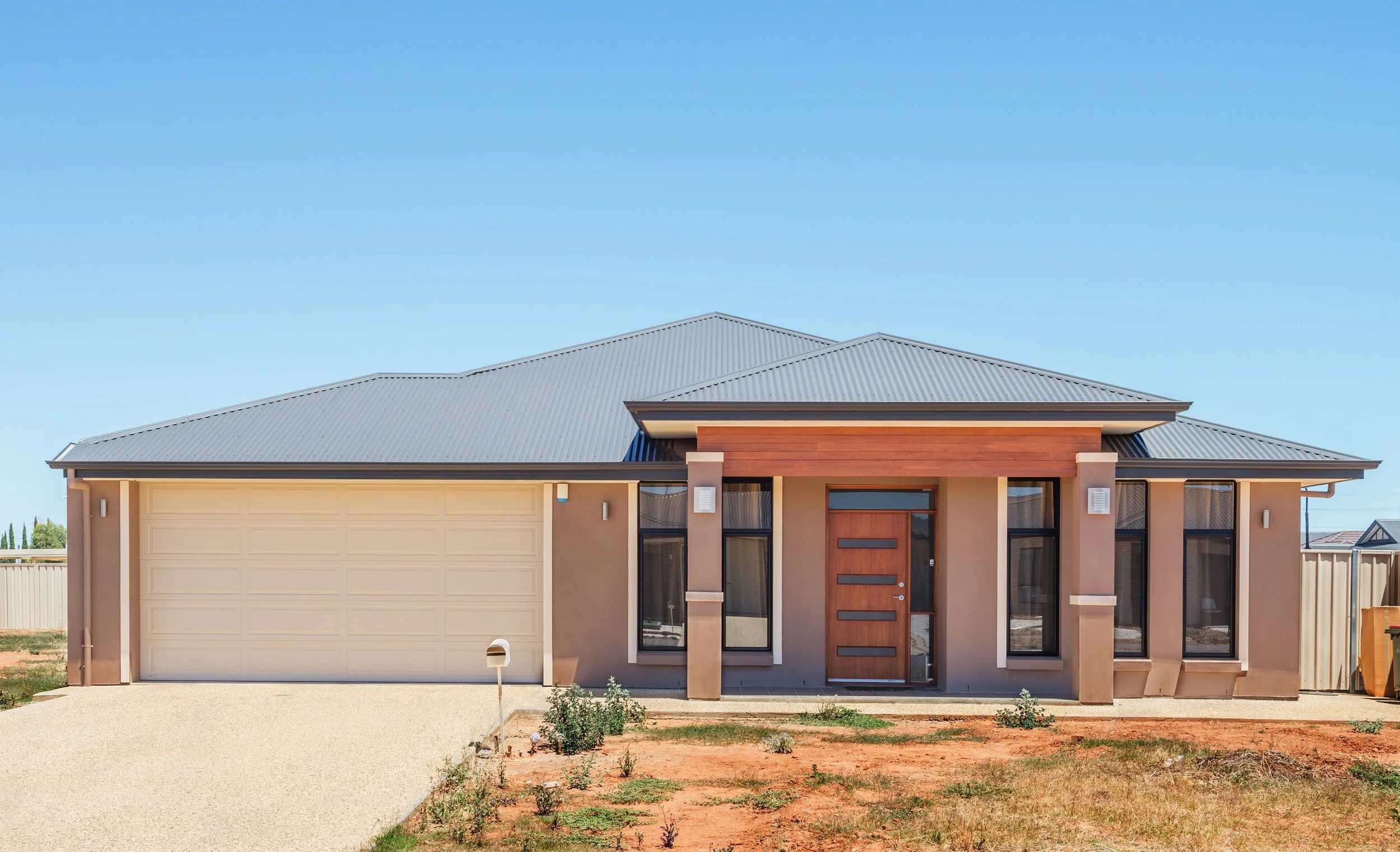 Modern single-story house with a beige garage door, a wooden front door, and large glass windows under a metal roof, set against a clear blue sky.
