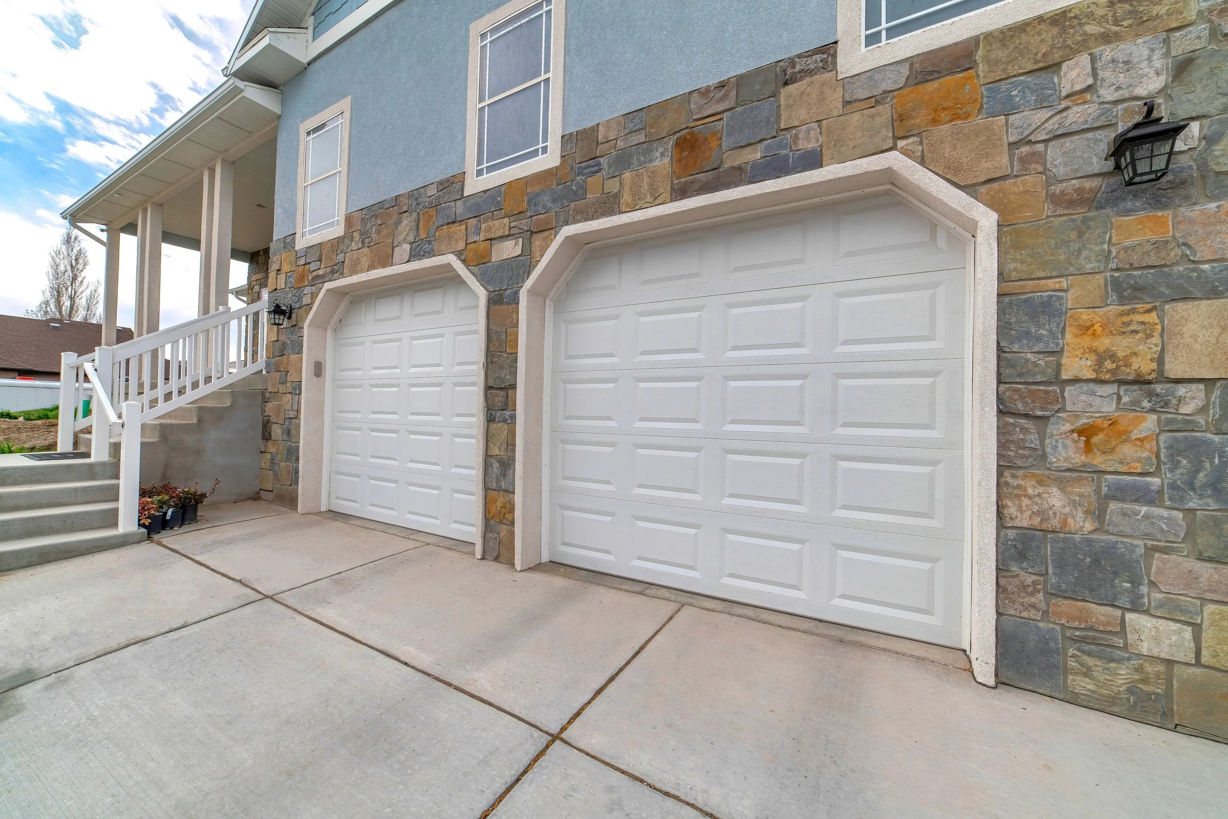 Image of a house with a stone and stucco exterior, two white garage doors, a staircase with white railings, and outdoor lights, under a partly cloudy sky.