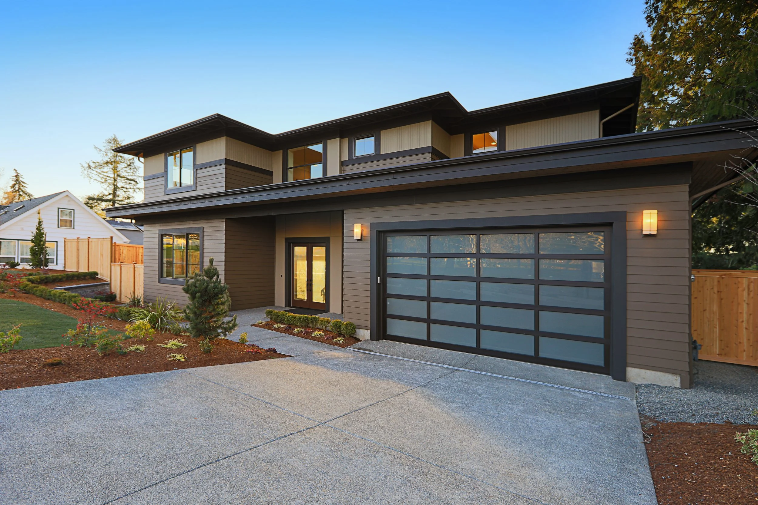 Modern two-story house with a gray garage door, front entrance with double glass doors, landscaped yard, and warm exterior lighting in the evening.