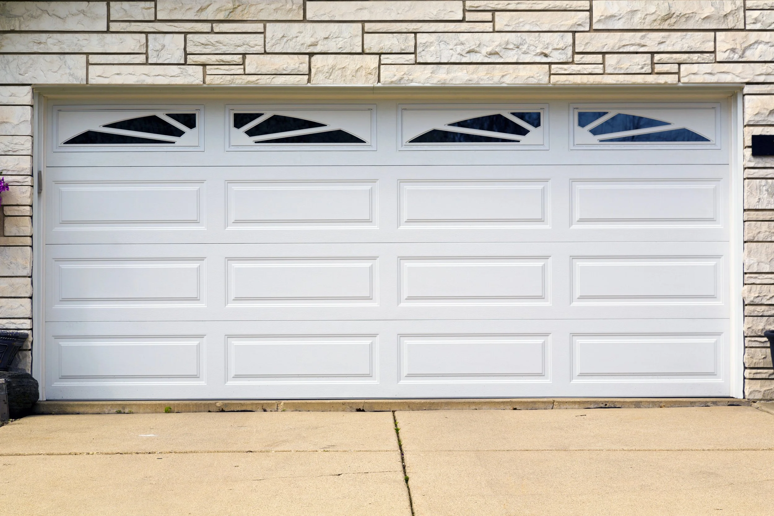 A white garage door with decorative windows at the top, set in a stone/brick house exterior, next to a sidewalk.