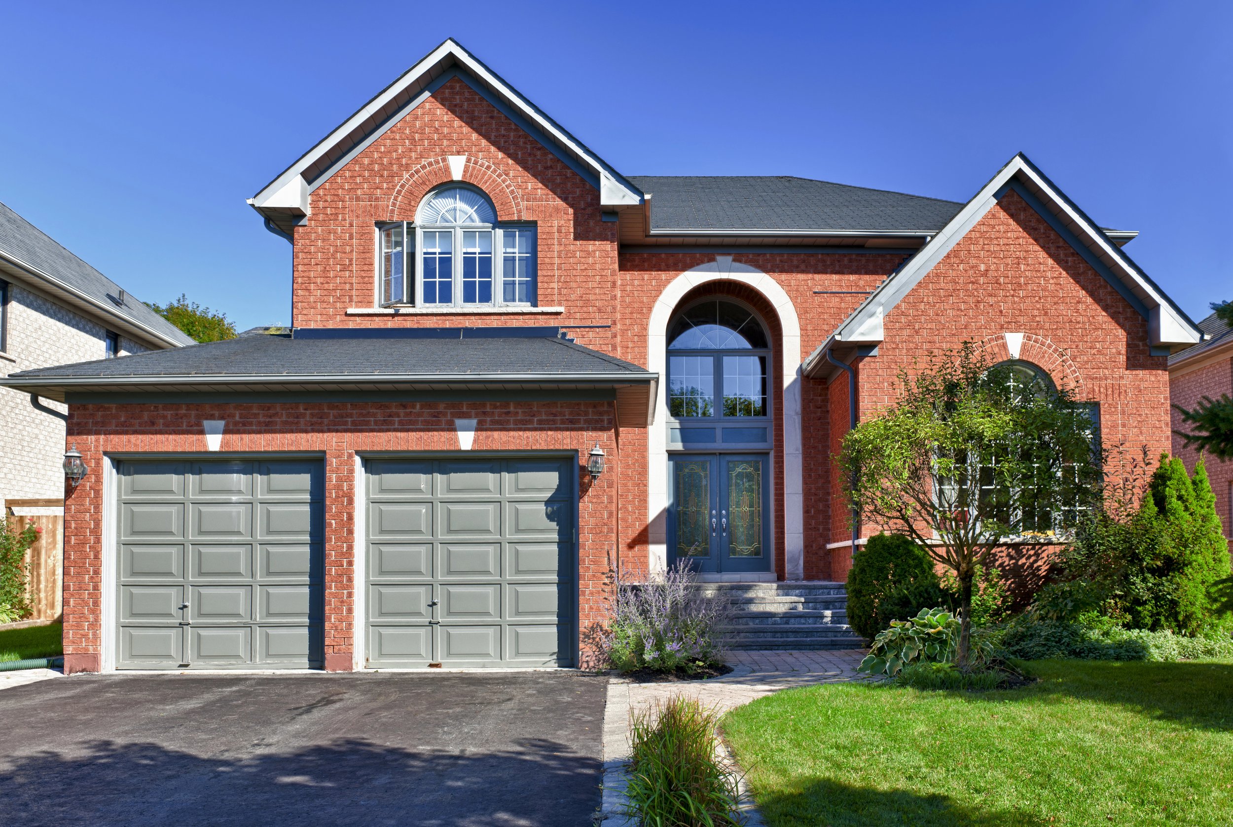 Two-story brick house with grey garage doors, a front door, driveway, and landscaped front yard containing a small tree and bushes, under a clear blue sky.