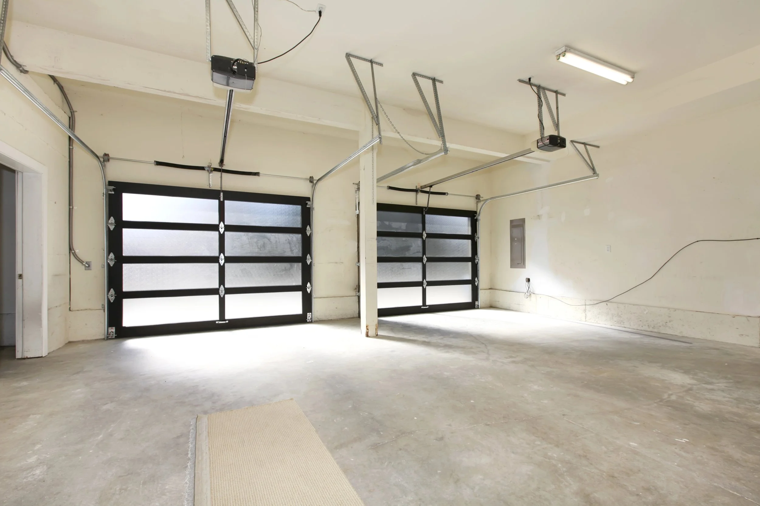 Empty garage with concrete floor, black garage doors, a mat on the floor, fluorescent ceiling light, and garage door openers mounted on the ceiling.
