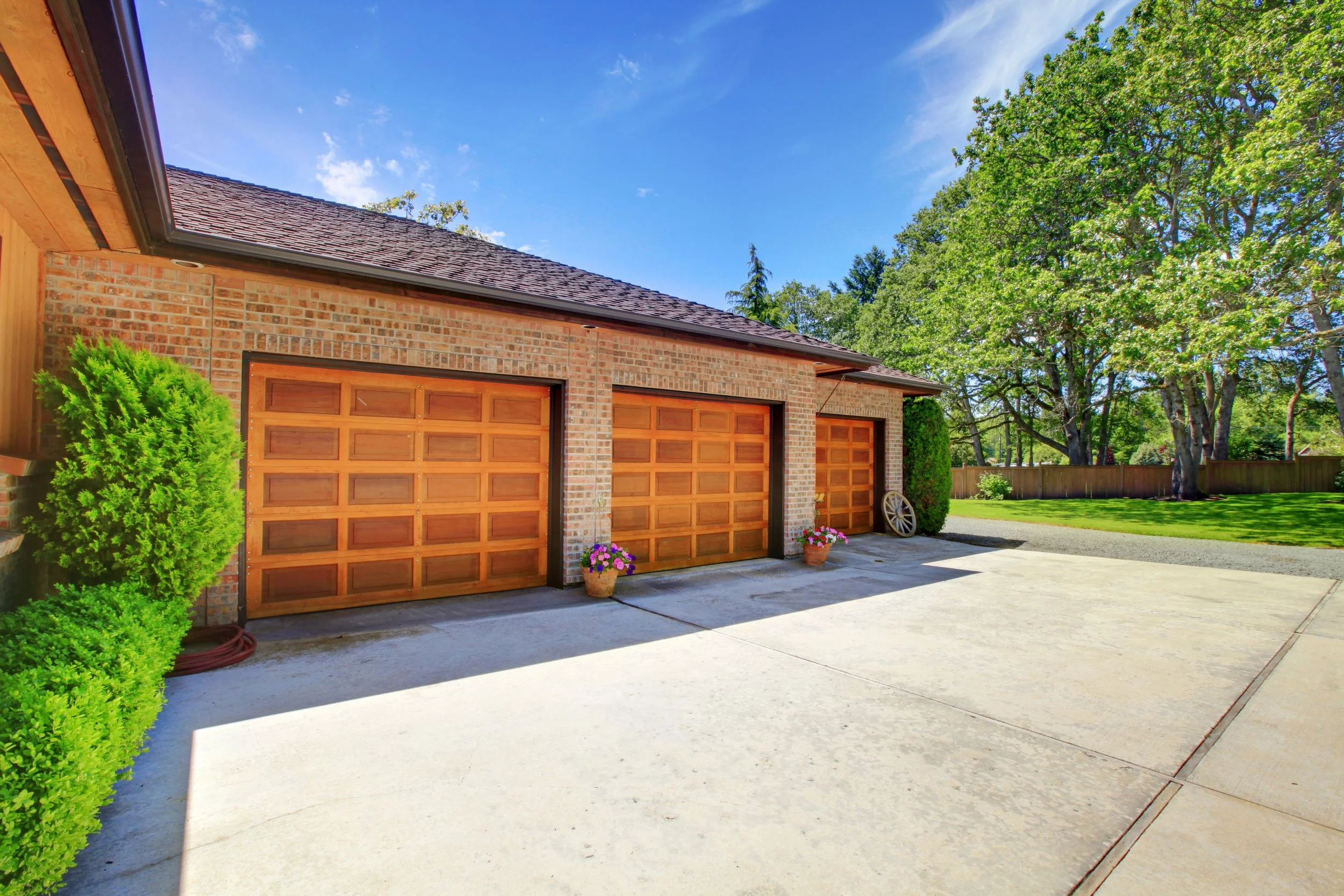 A residential driveway with two wooden garage doors, a brick house exterior, a tree, potted flowers, and a clear blue sky.