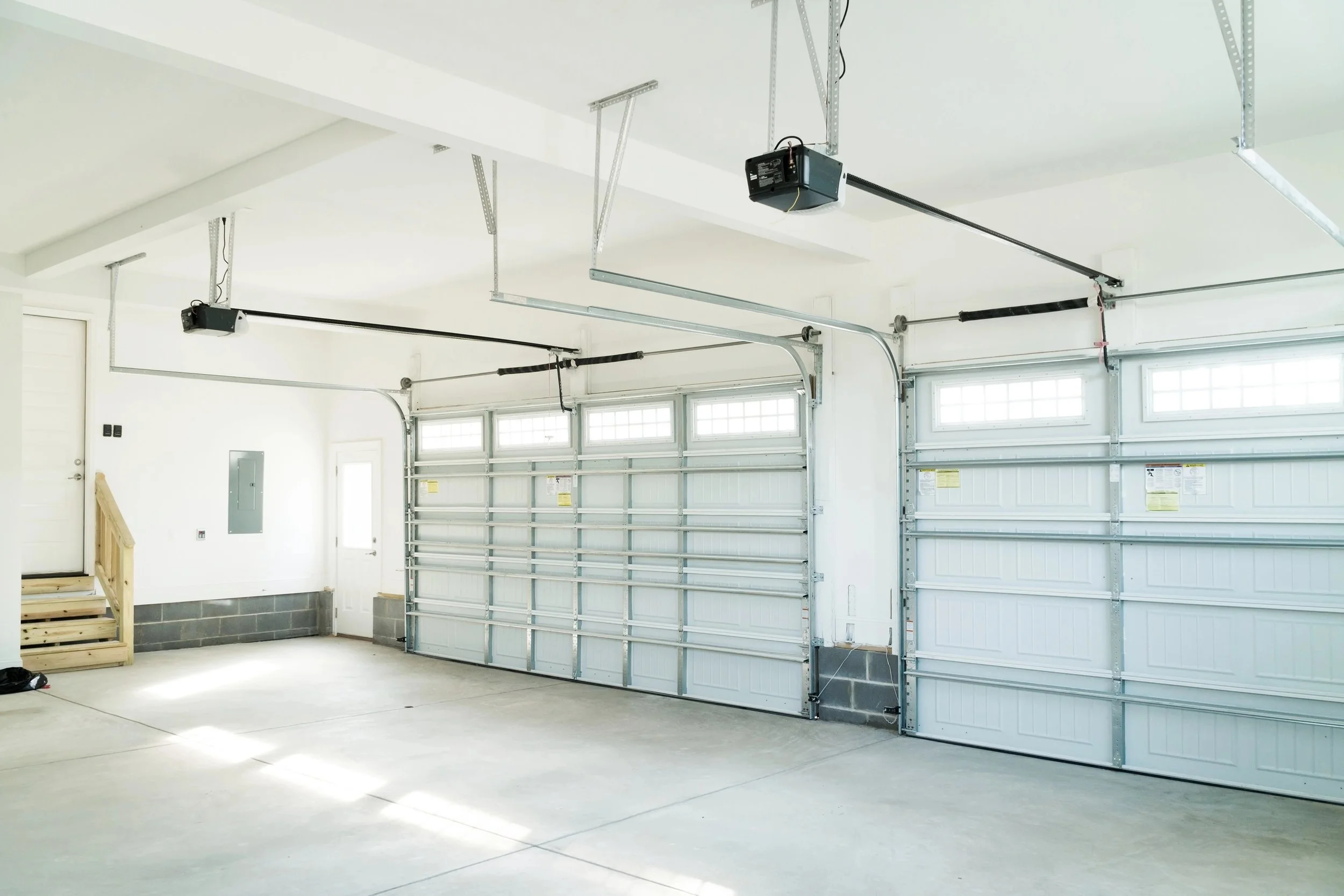 Empty two-car garage with gray steel garage doors, concrete floor, and windows above the doors. Open garage door mechanism with metal tracks and motors visible on the ceiling. Small stairs on the left side leading to an elevated entryway.