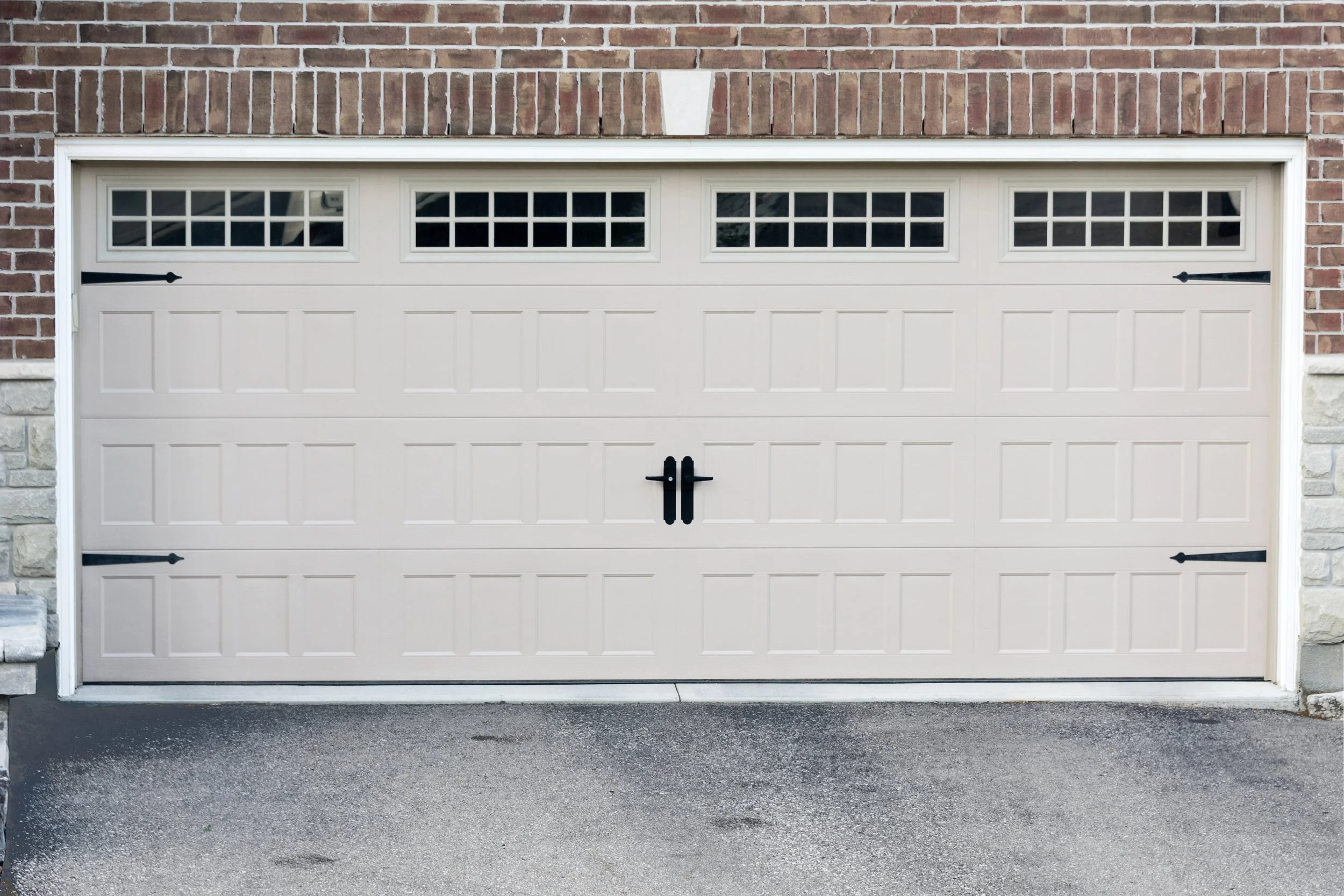 White garage door with small window panels at the top, set in a brick wall with stone accents on the sides, and black hardware, situated above asphalt driveway.