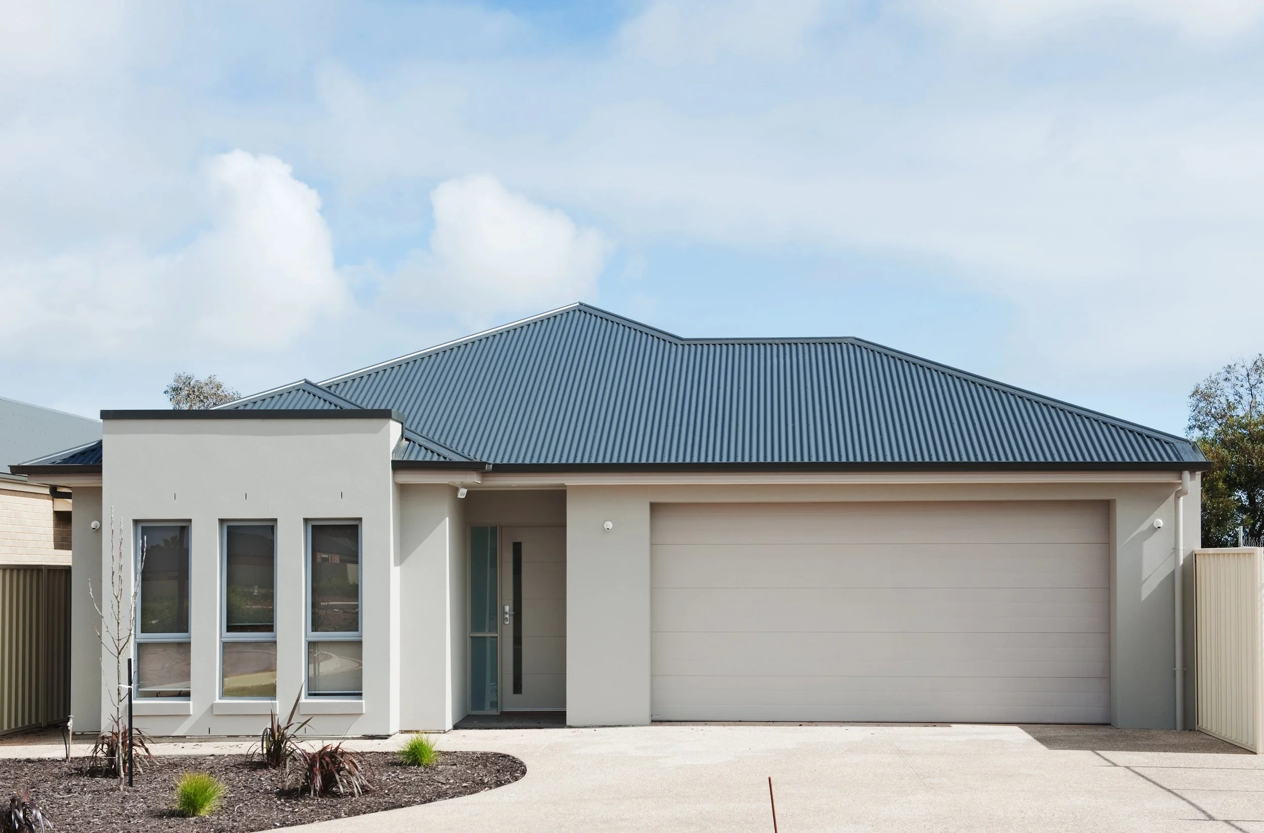 Modern house with white walls and a metal roof, front yard with small plants, and a cloudy sky.