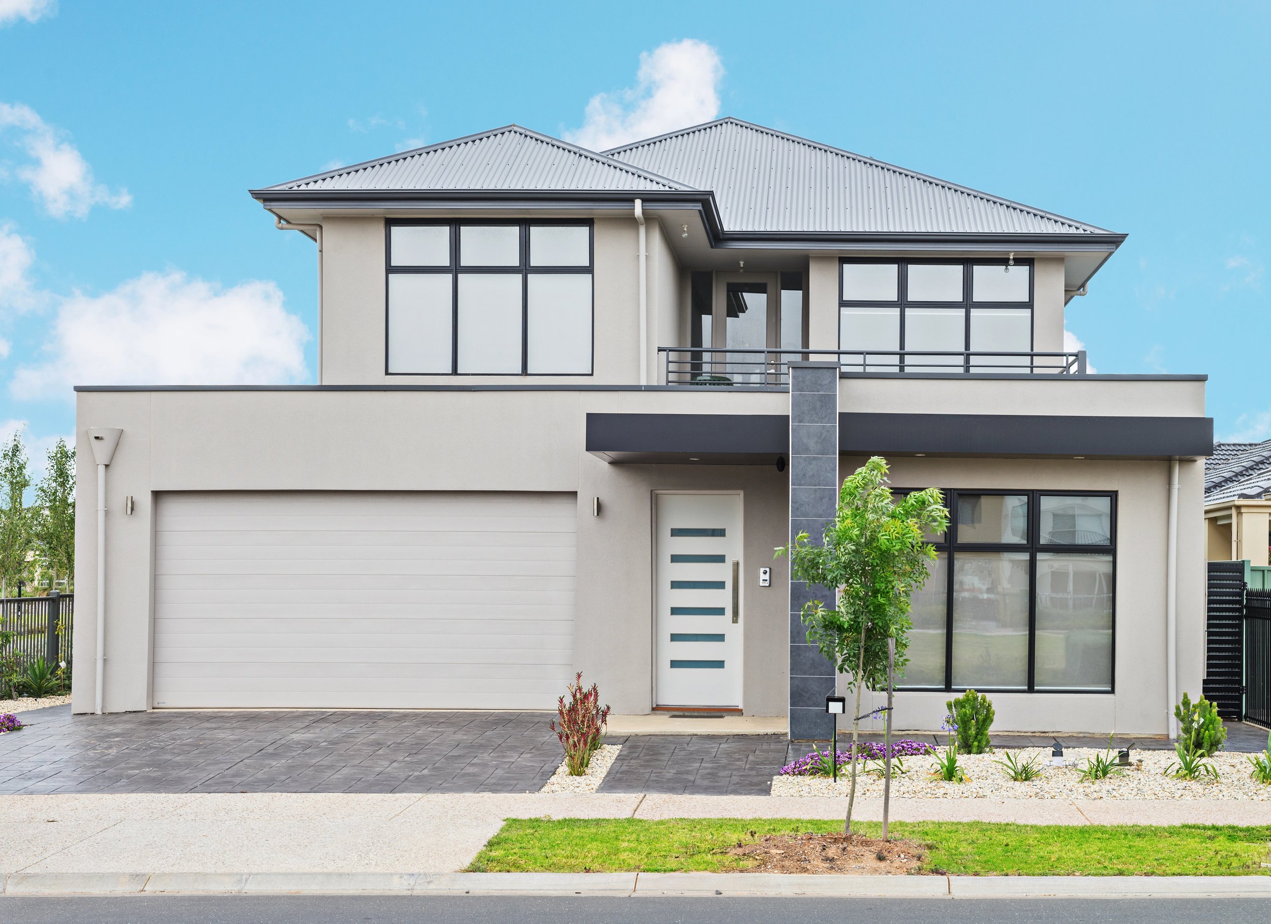 Modern two-story house with a gray metal roof, large windows, white walls, and a peaked roof design. The front yard has a small tree, bushes, and a garden with purple flowers, with a paved driveway and sidewalk.