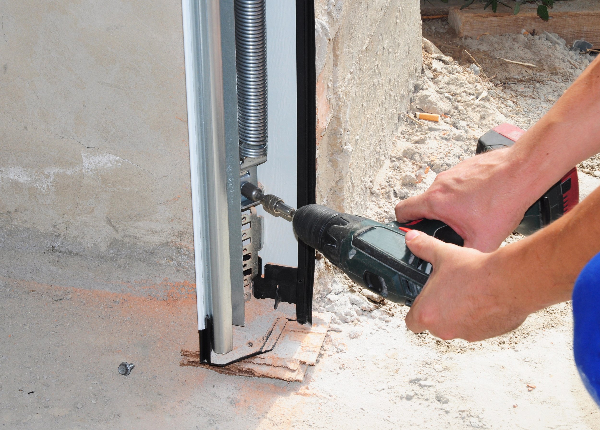 A person using a power drill to install or repair a metal door frame on a construction site with dirt and construction materials around.