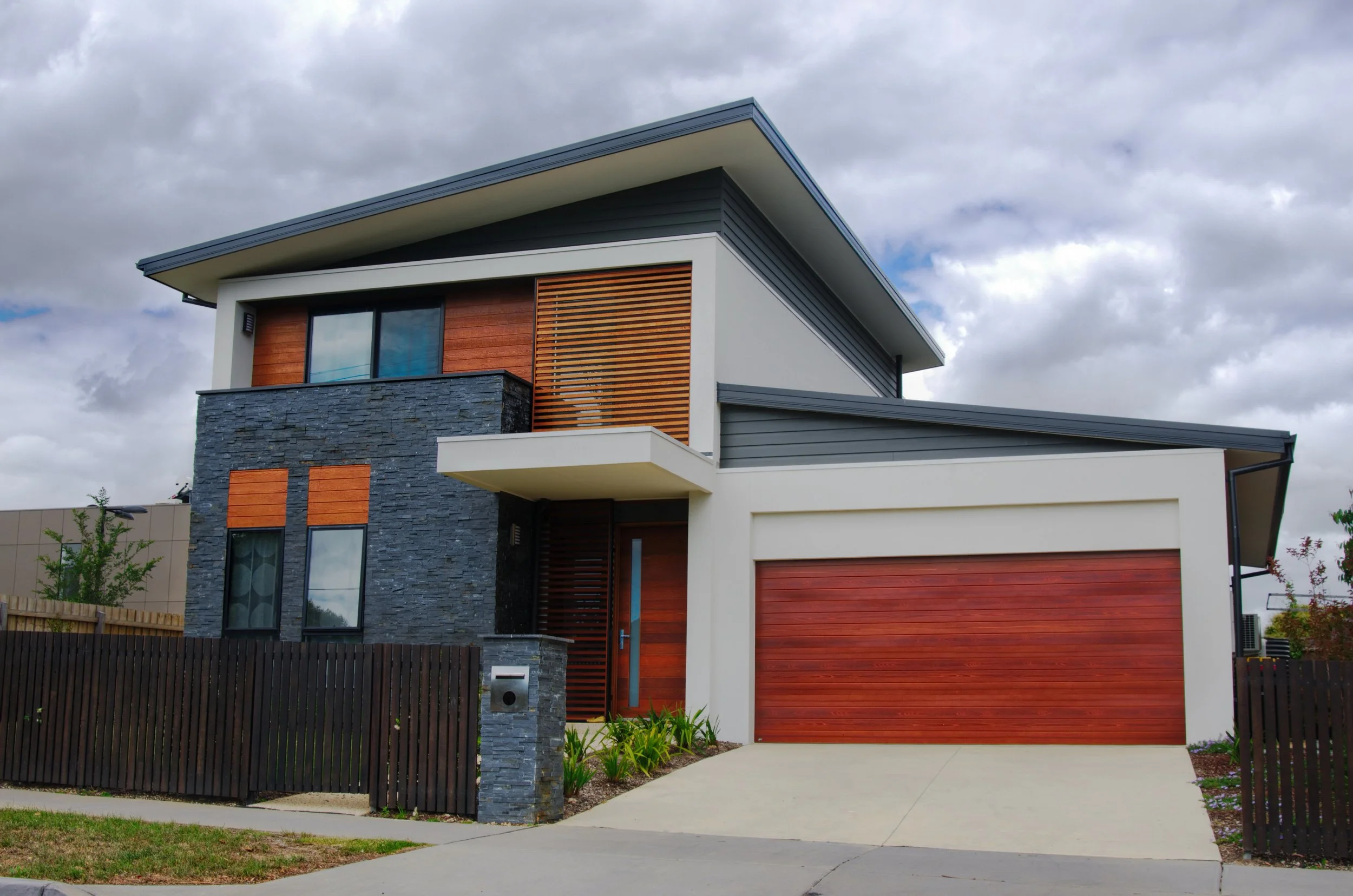 Modern two-story house with mixed materials, including stone, wood, and metal siding, featuring a garage door, a balcony, and landscaped yard, under cloudy sky.