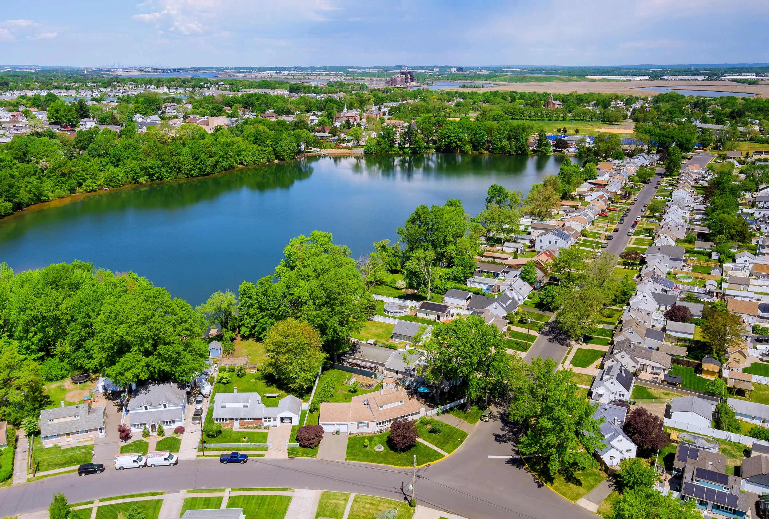 Aerial view of a suburban neighborhood with a lake, surrounded by trees, houses, and streets, with a city skyline and industrial area in the distance.