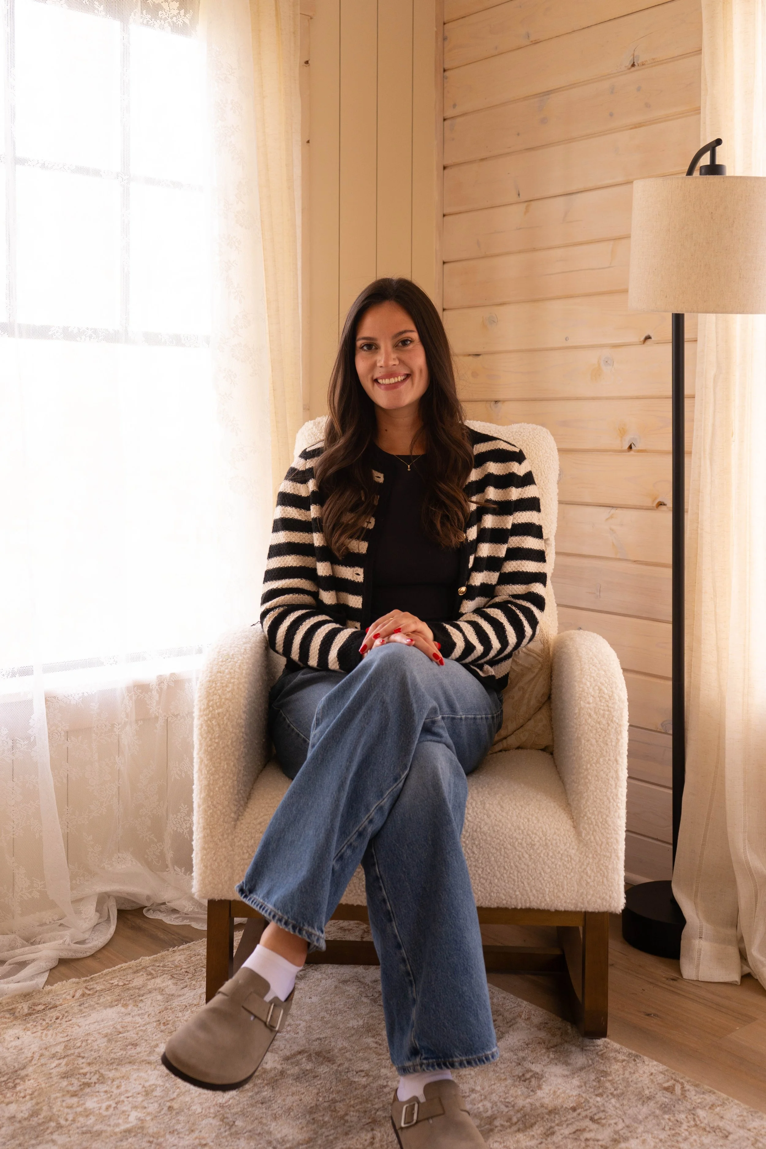 A woman sitting in a cream-colored armchair in a cozy room with wooden walls and a large window with sheer curtains. She is smiling, wearing a black top, a black and white striped cardigan, blue jeans, beige slip-on shoes, and white socks.