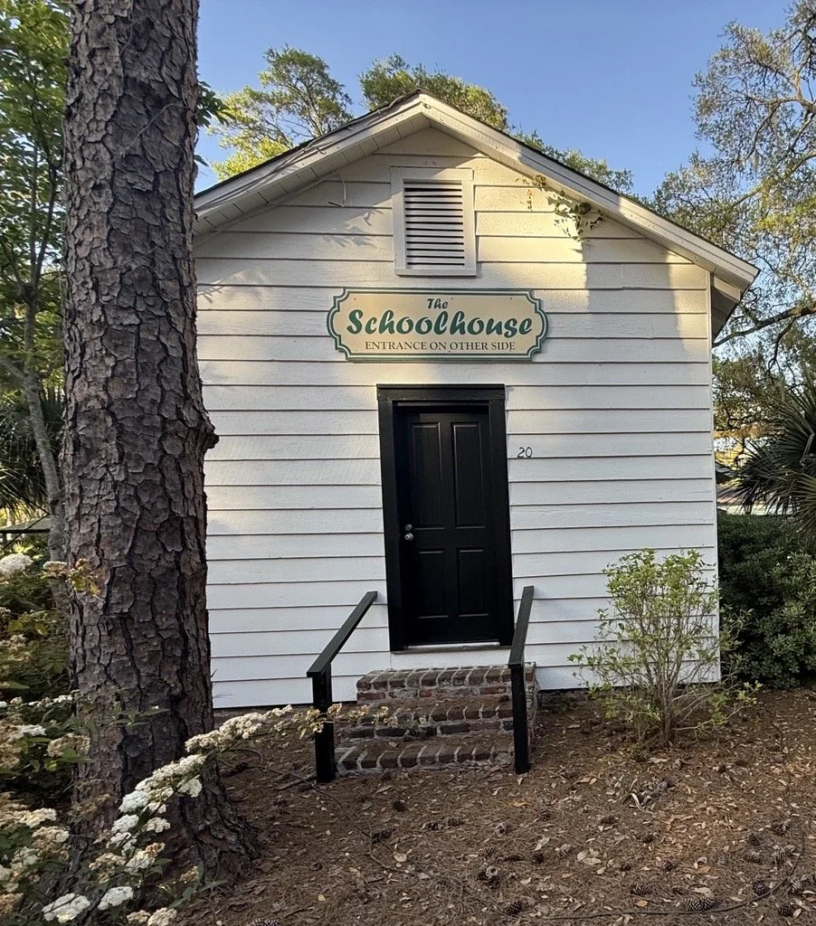 A small white wooden building with a gable roof, labeled 'The Schoolhouse' with a sign indicating an alternate entrance. It has a black door, brick steps, and is surrounded by trees and bushes.
