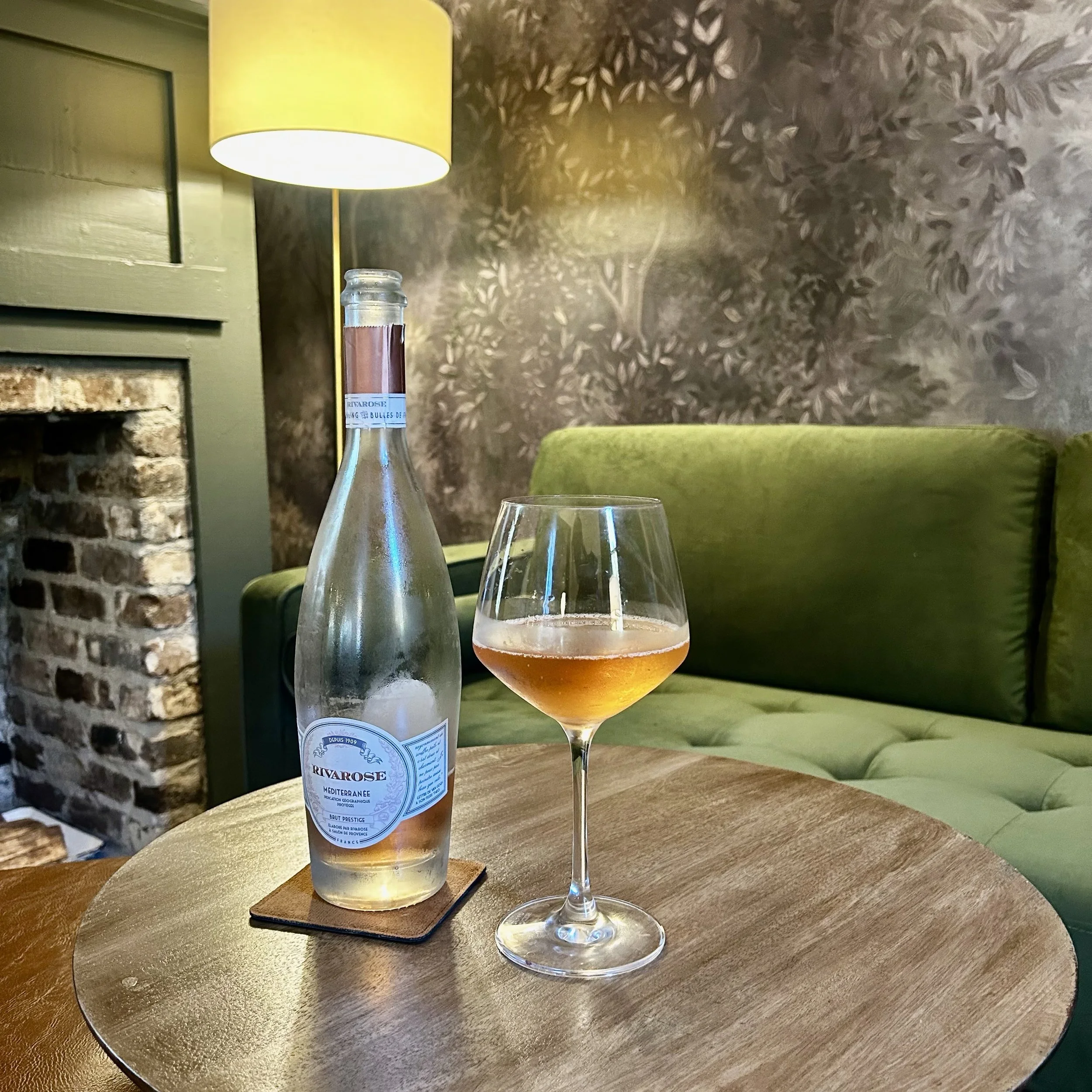 A clear glass bottle of white wine with a blue and white label, a filled wine glass, a table, a green upholstered chair, a yellow lamp, a textured brown and beige wall, and part of a brick fireplace in a cozy restaurant or cafe setting.