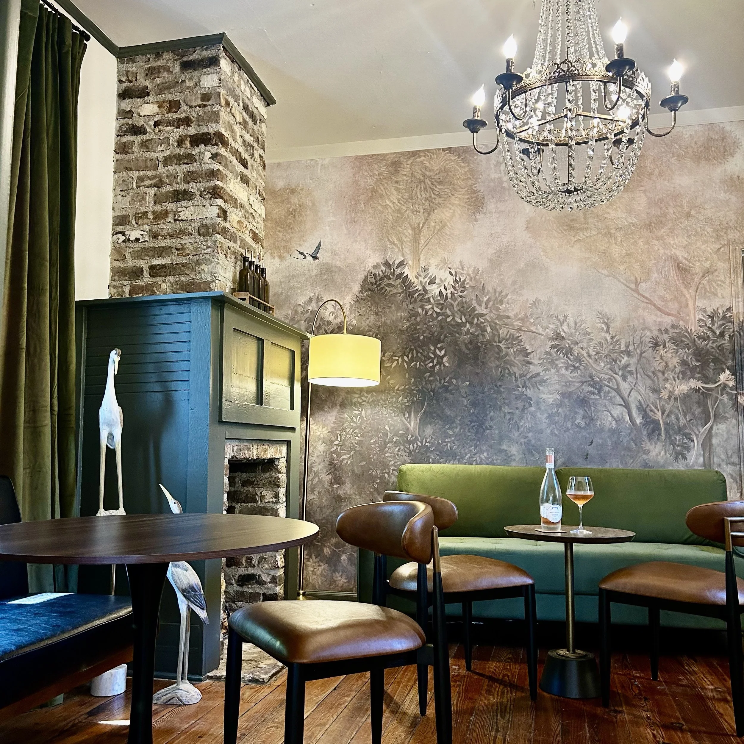 Interior of a restaurant or cafe with a brick wall and large chandelier, green curtain, wooden floors, wooden chairs, a table with a wine glass and a bottle of water.