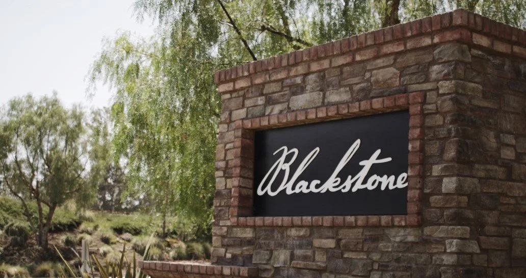A brick sign with the word "Blackstone" written in cursive, surrounded by trees and greenery.