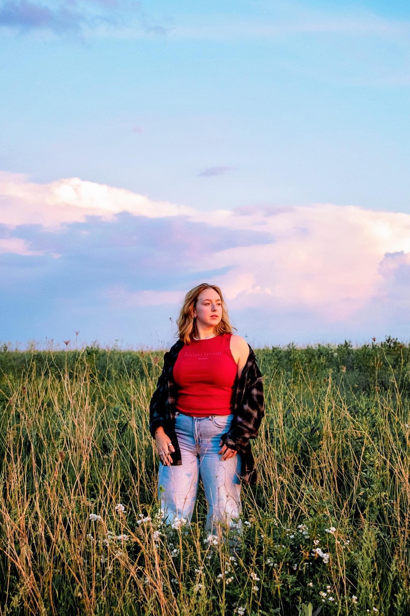 A young woman standing in a grassy field with wildflowers, wearing a red sleeveless top, light jeans, and a black and red plaid shirt, with the sky showing pink and purple clouds at sunset.