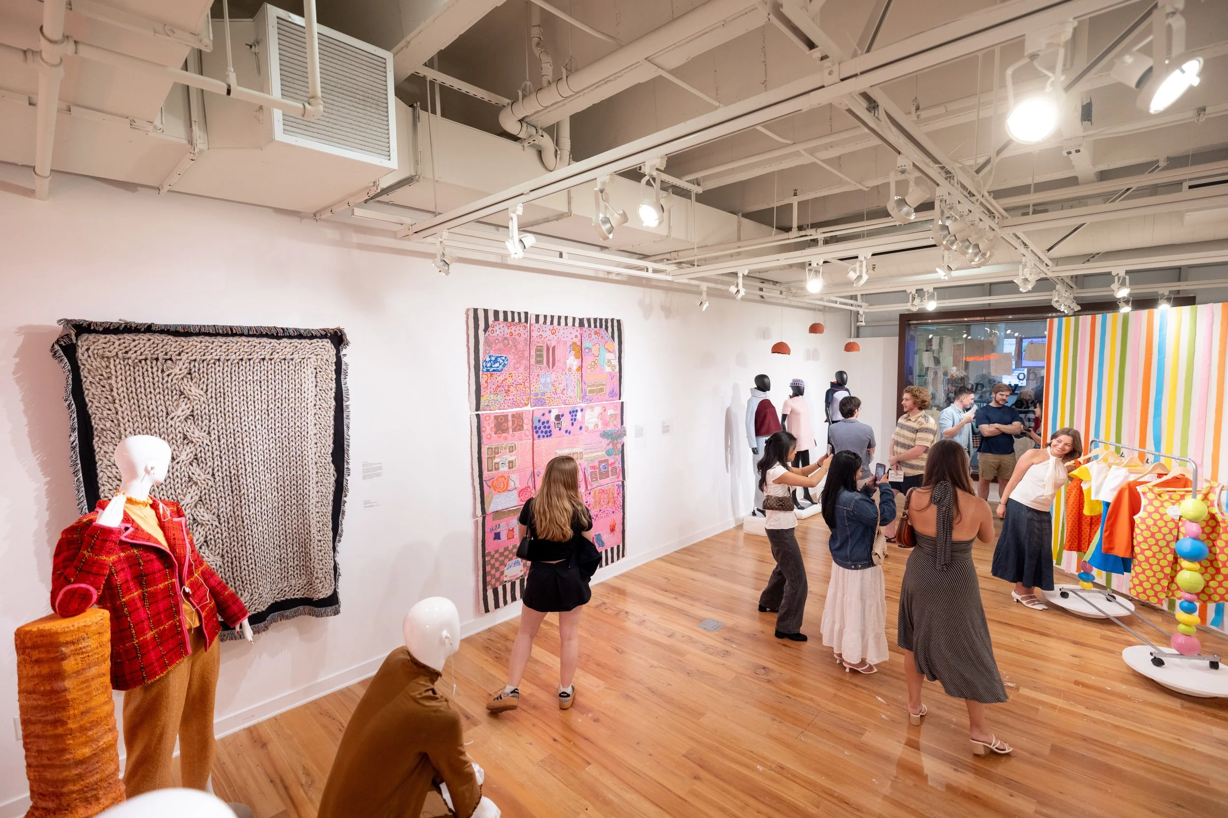 People visiting an art gallery or exhibit, viewing colorful artwork and mannequins dressed in clothing. Some visitors are taking photos, while others are engaging with the displays and chatting.