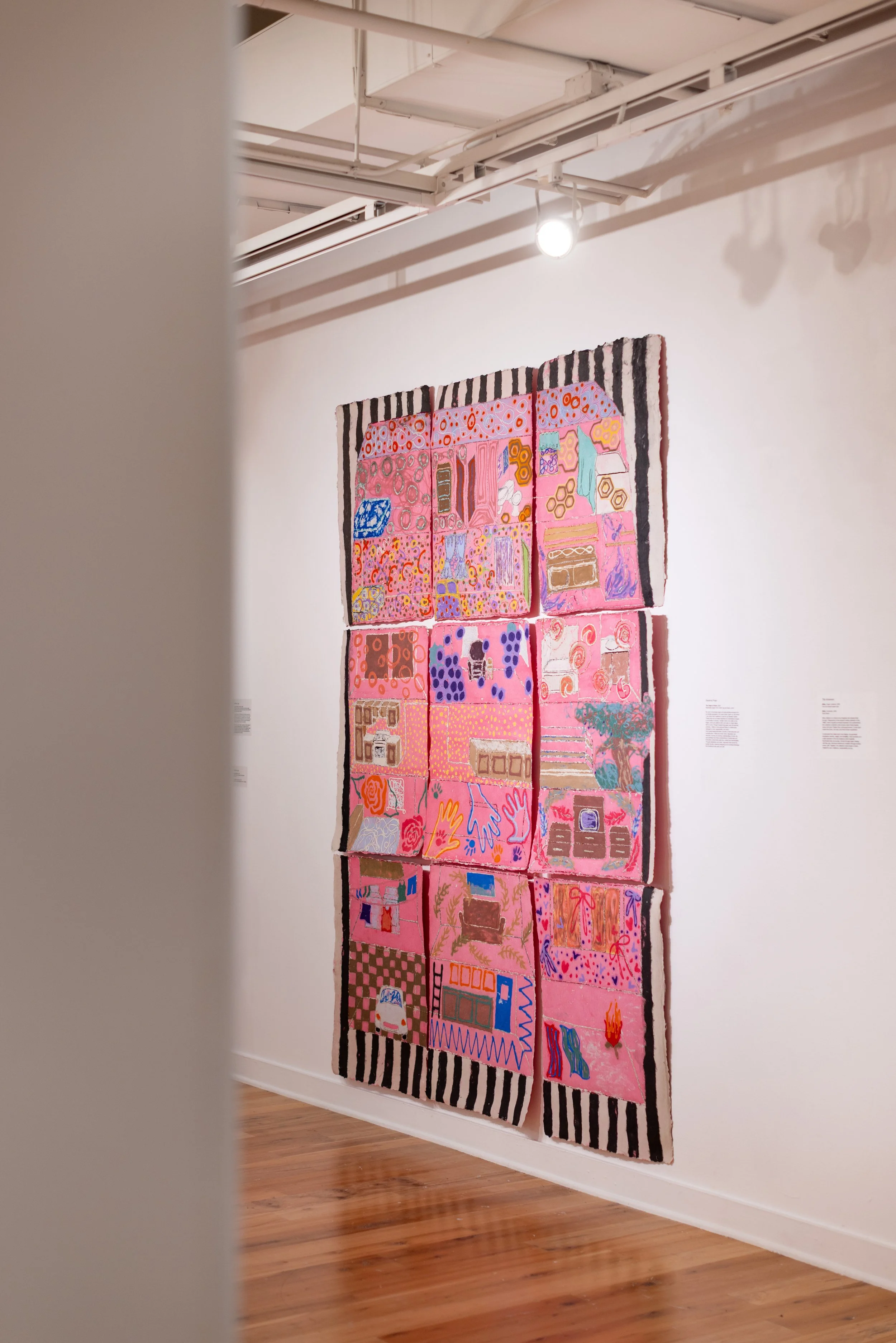 Colorful, patterned fabric artwork displayed on a white gallery wall, with a black and white striped border at the top and bottom, and a light fixture overhead illuminating the piece.