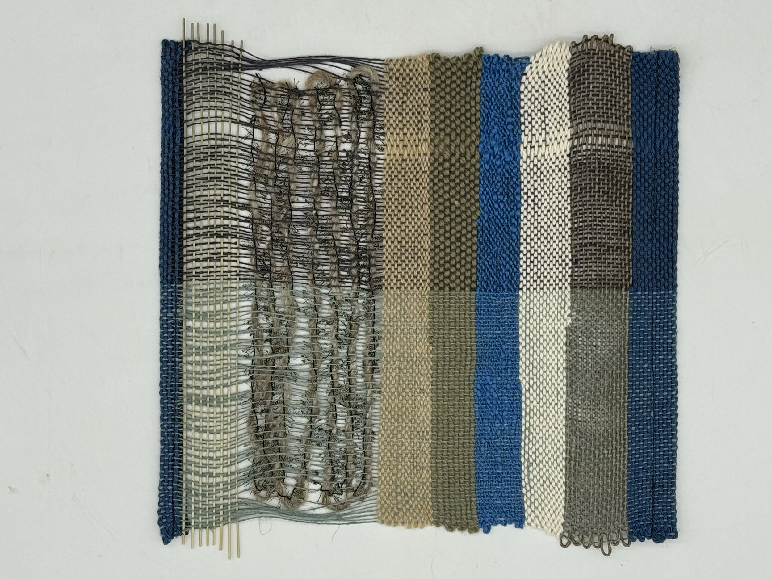 A textile art piece made of woven fabric strips in various colors and patterns, mounted on a wall with a section of metal pins at the top.