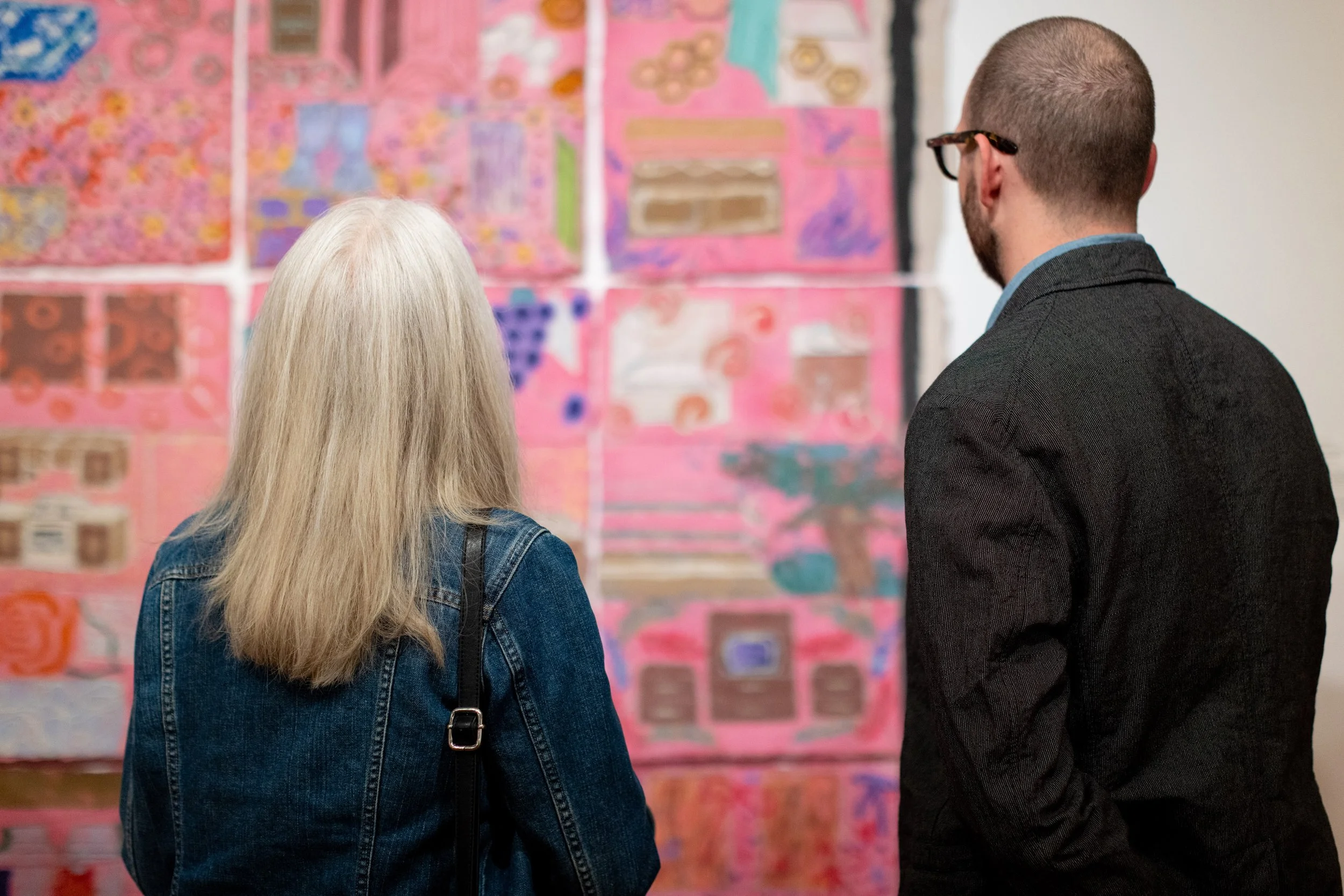 Two people, one with long white hair and a denim jacket and the other with short hair and wearing glasses and a dark jacket, look at colorful abstract artwork on a wall in an art gallery.
