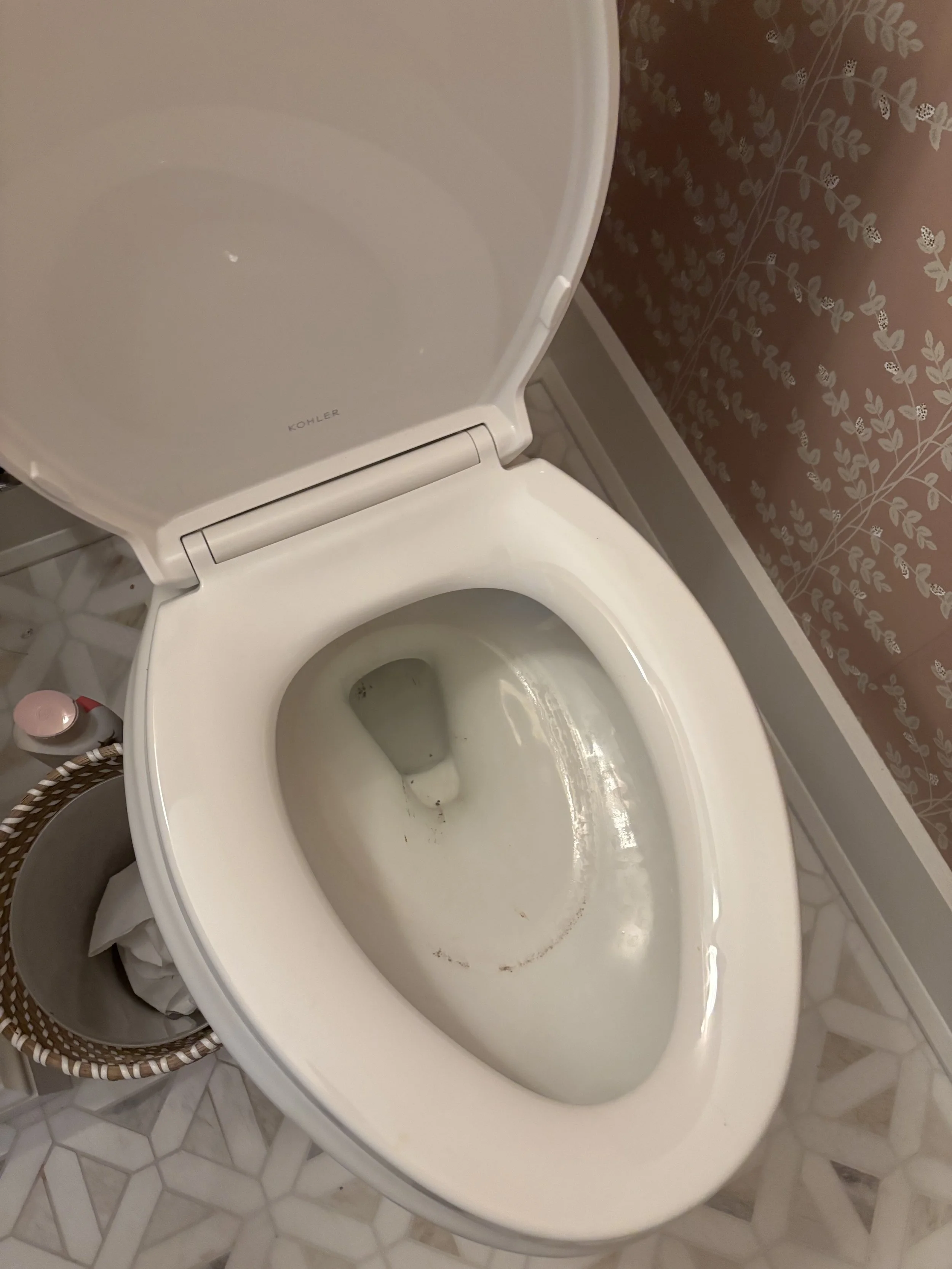 Empty toilet with a slightly dirty bowl, a basket with paper towels and a bottle of toilet cleaner on the side, patterned tile flooring, and floral wallpaper in the background.