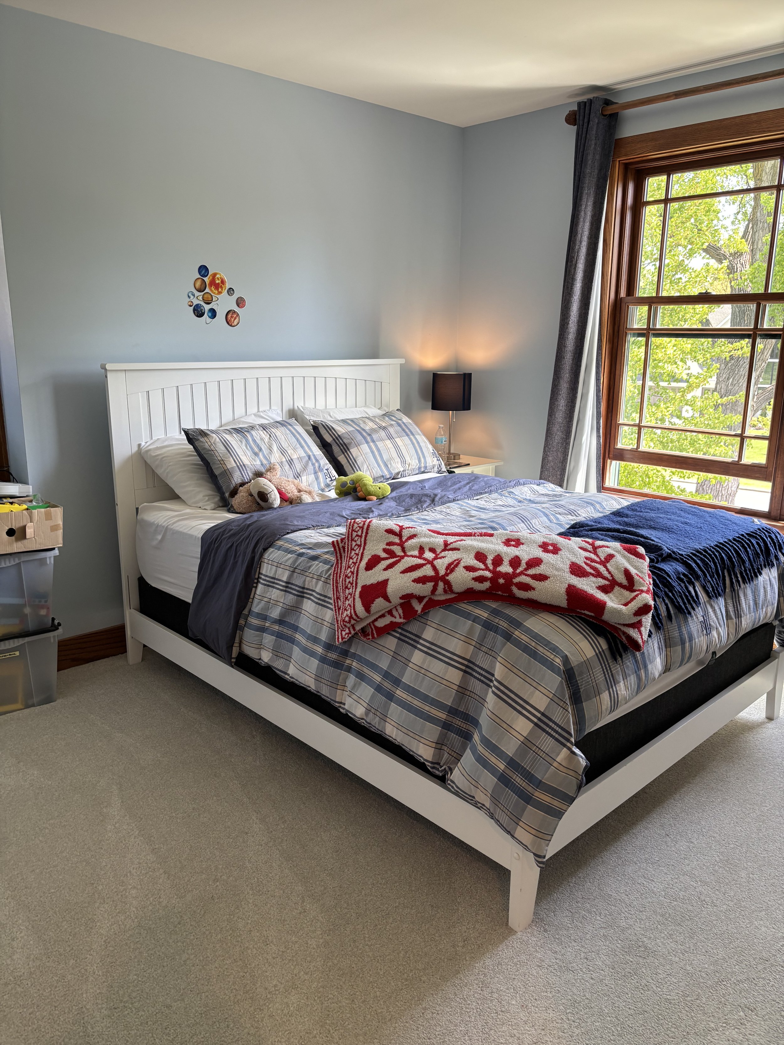 A bedroom with a white wooden bed frame, a plaid bedspread, stuffed animals, a nightstand with a lamp, a window with a tree outside, and a blue and red patterned blanket on the bed.