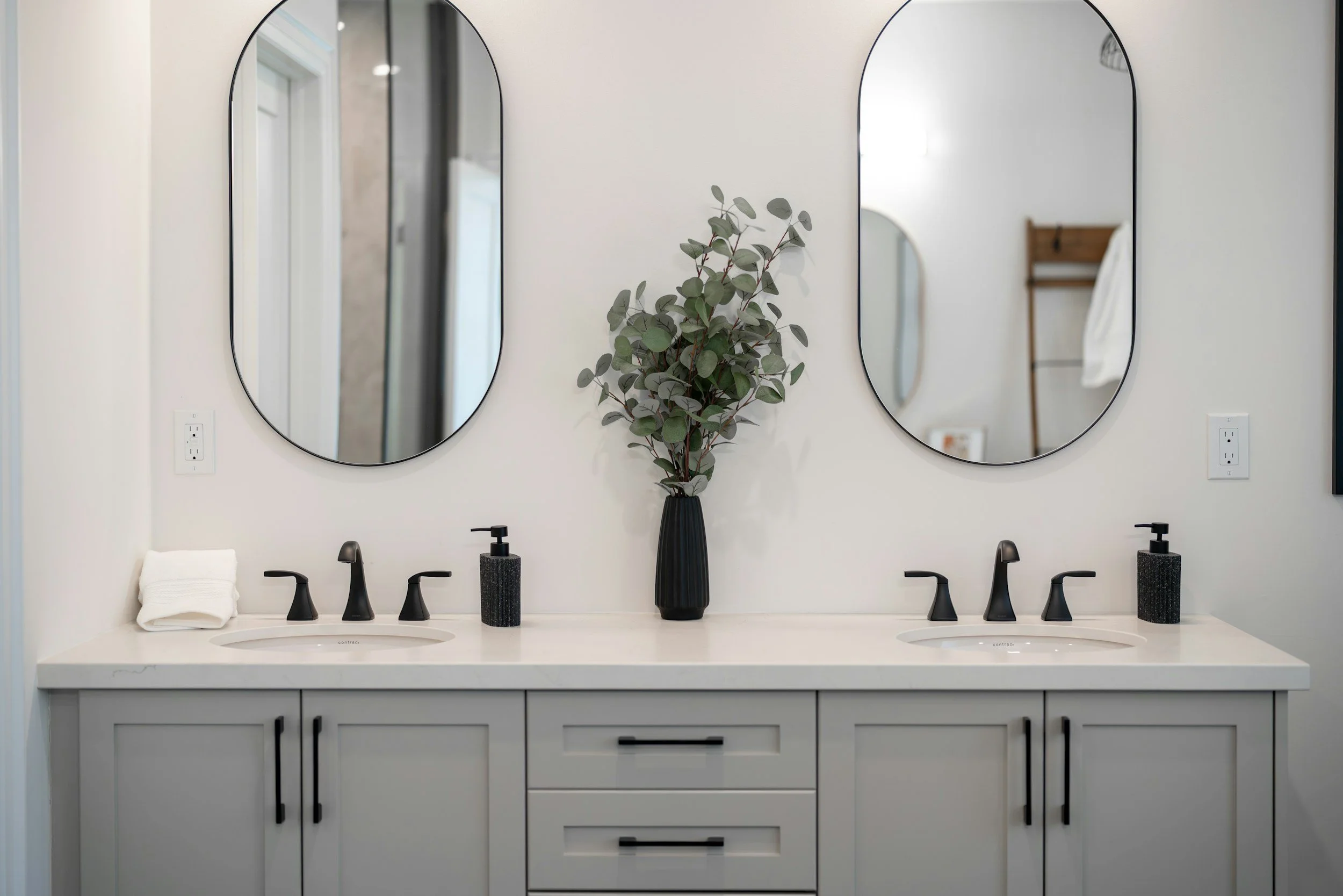 A modern bathroom vanity with two white sinks, black faucets, and two oval mirrors on a white wall. There are black soap dispensers, a neatly folded white towel, a black vase with green leaves, and a wooden towel rack with a white towel.