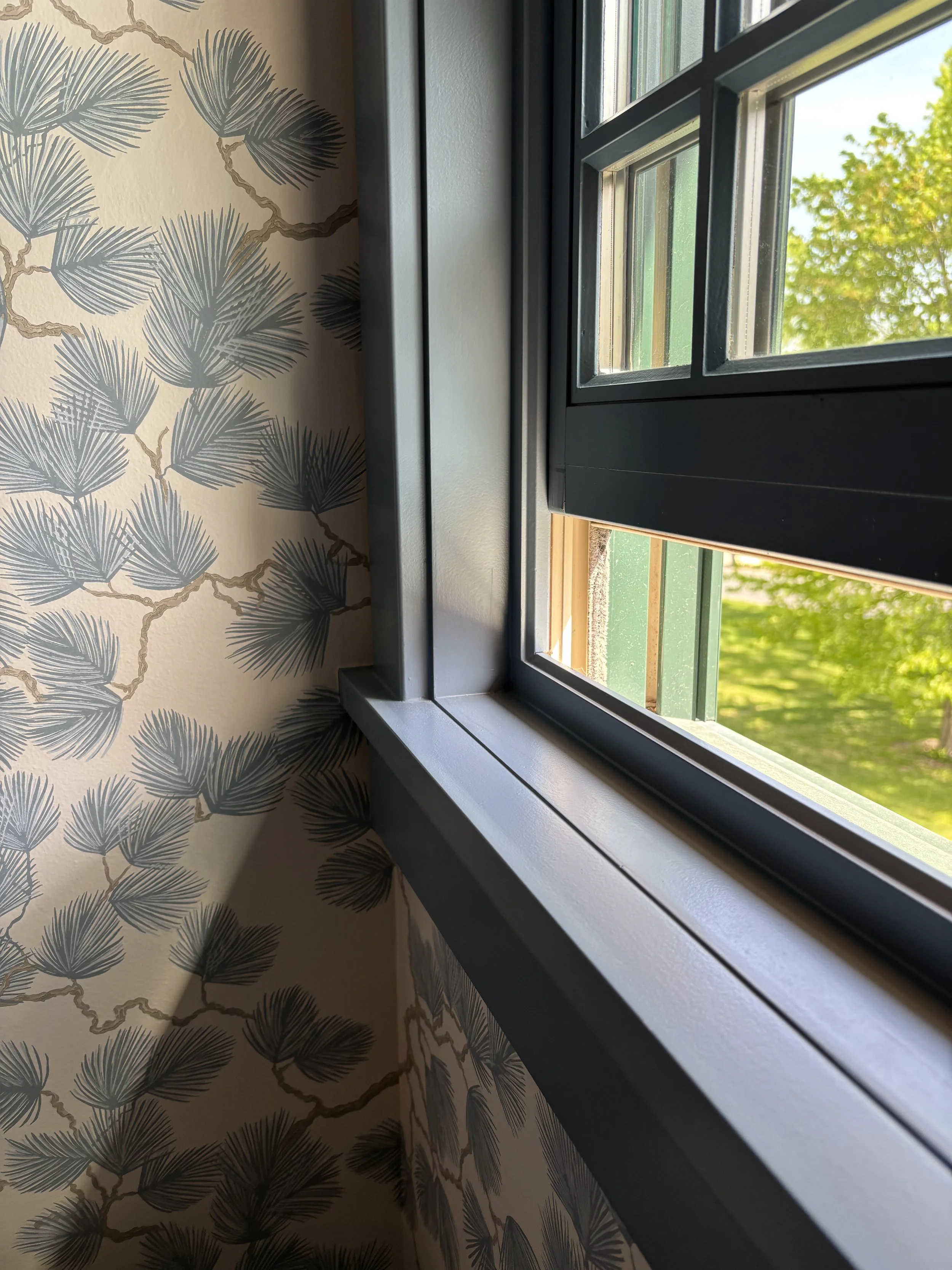 View of a window with black framing, looking outside at green trees and a blue sky, with wallpaper featuring a pattern of dark blue pine leaves with gold outlines inside the room.