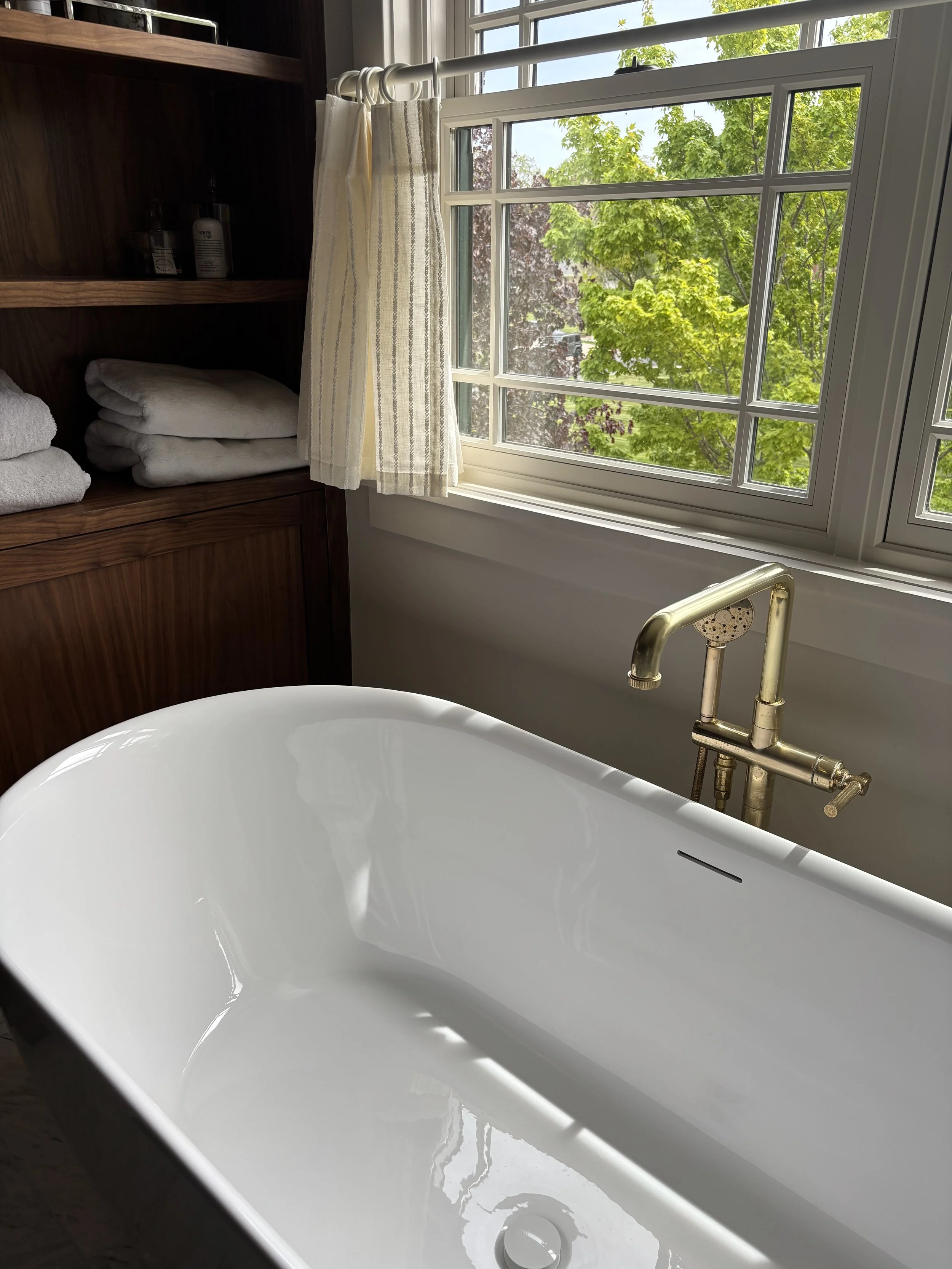 A white freestanding bathtub with a brass faucet located next to a large window showing leafy trees outside, and a wooden cabinet with folded towels and personal care items nearby.