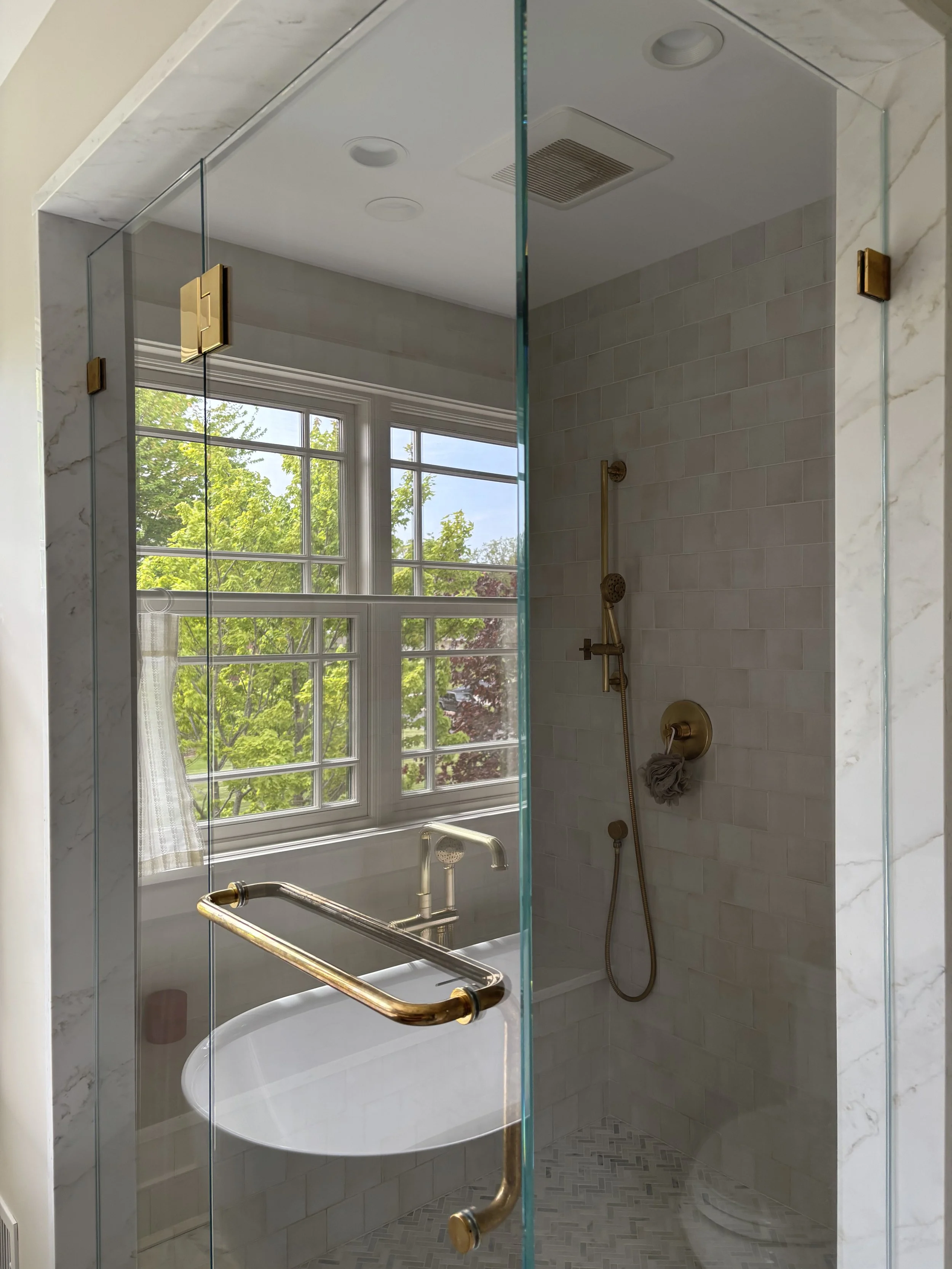 A bathroom shower with a glass door, featuring a large window with a view of green trees outside. The shower has a brass grab bar, a handheld showerhead, and a wall-mounted shower control. The interior is tiled with light-colored subway tiles and has