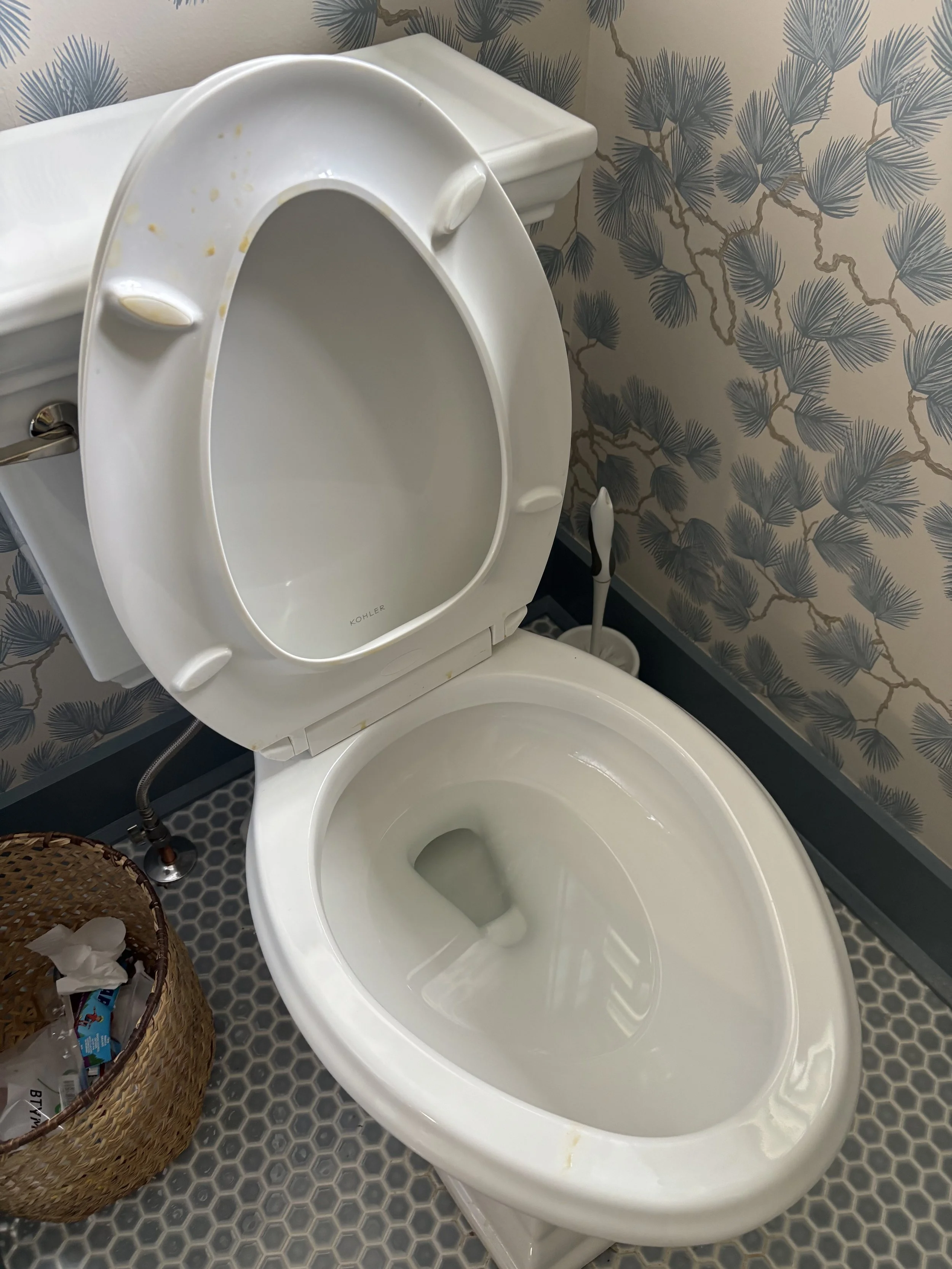 Empty white toilet with the lid up in a bathroom with patterned wallpaper and a hexagonal tile floor. There is a woven basket with tissues and some toilet paper on the side.