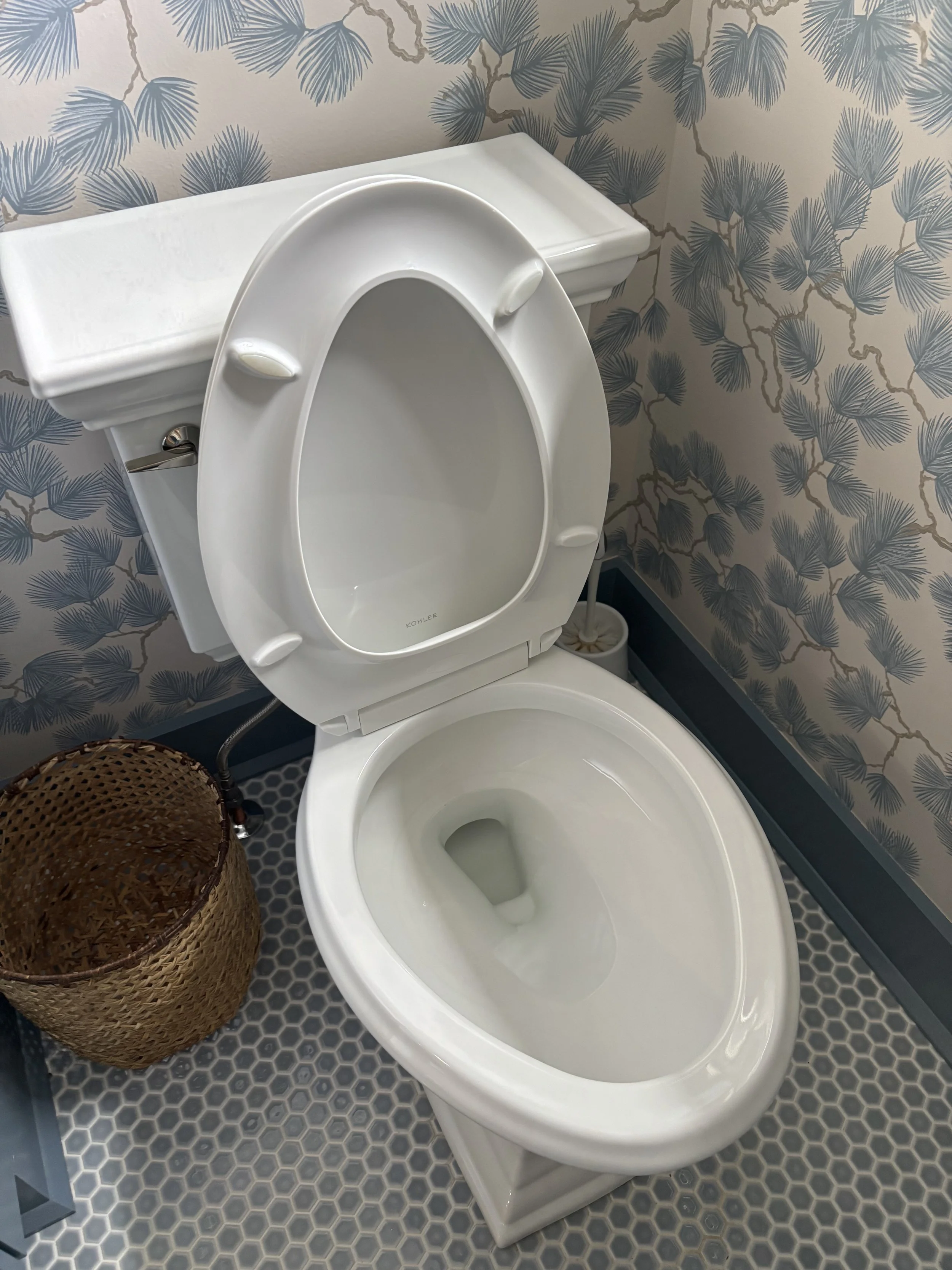 Empty white toilet bowl with the lid open, located in a bathroom with blue and beige leaf-patterned wallpaper and a honeycomb-patterned floor, next to a woven basket.
