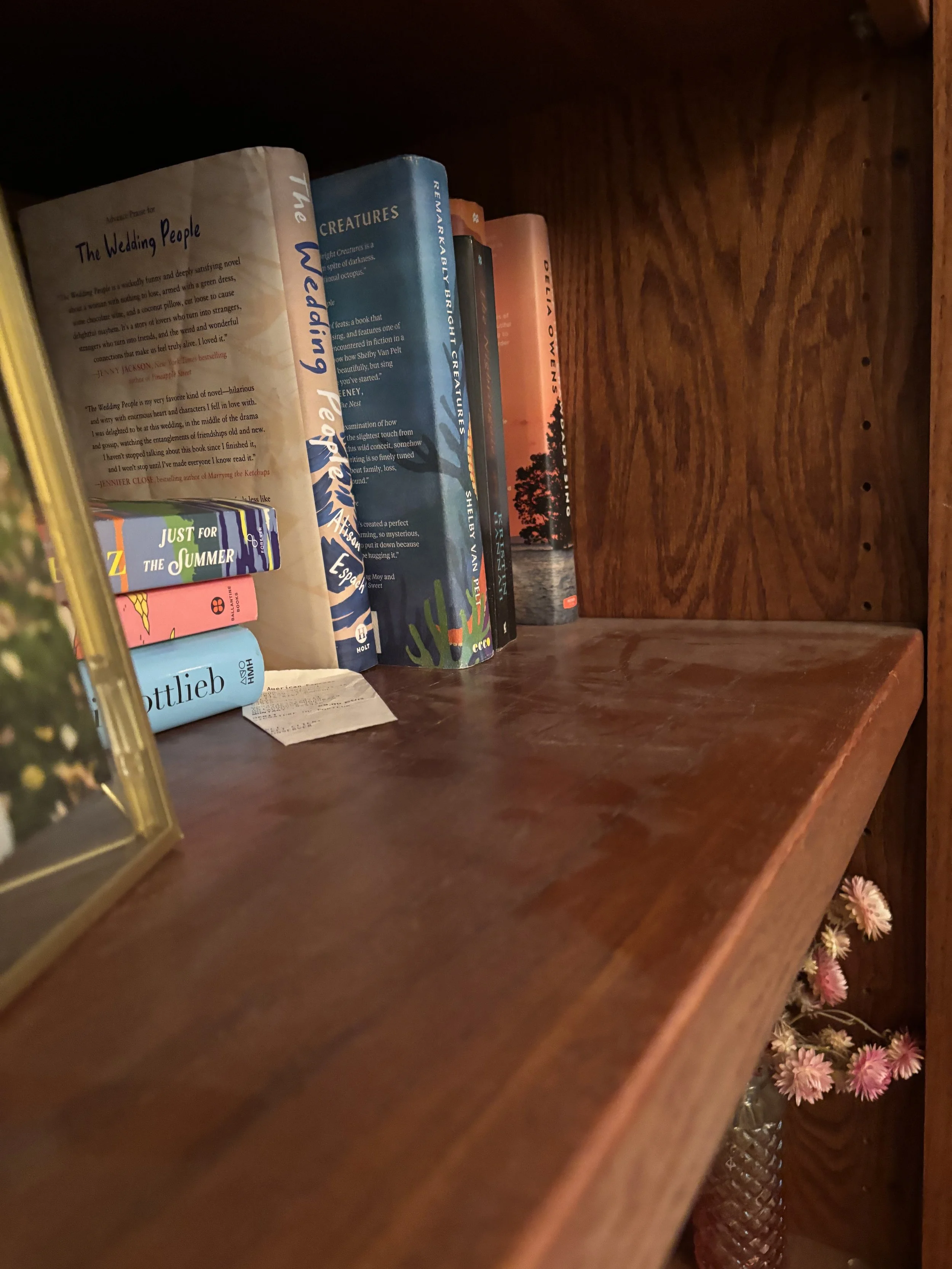 A wooden bookshelf holding various books, with pink flowers in a vase visible at the bottom right corner.