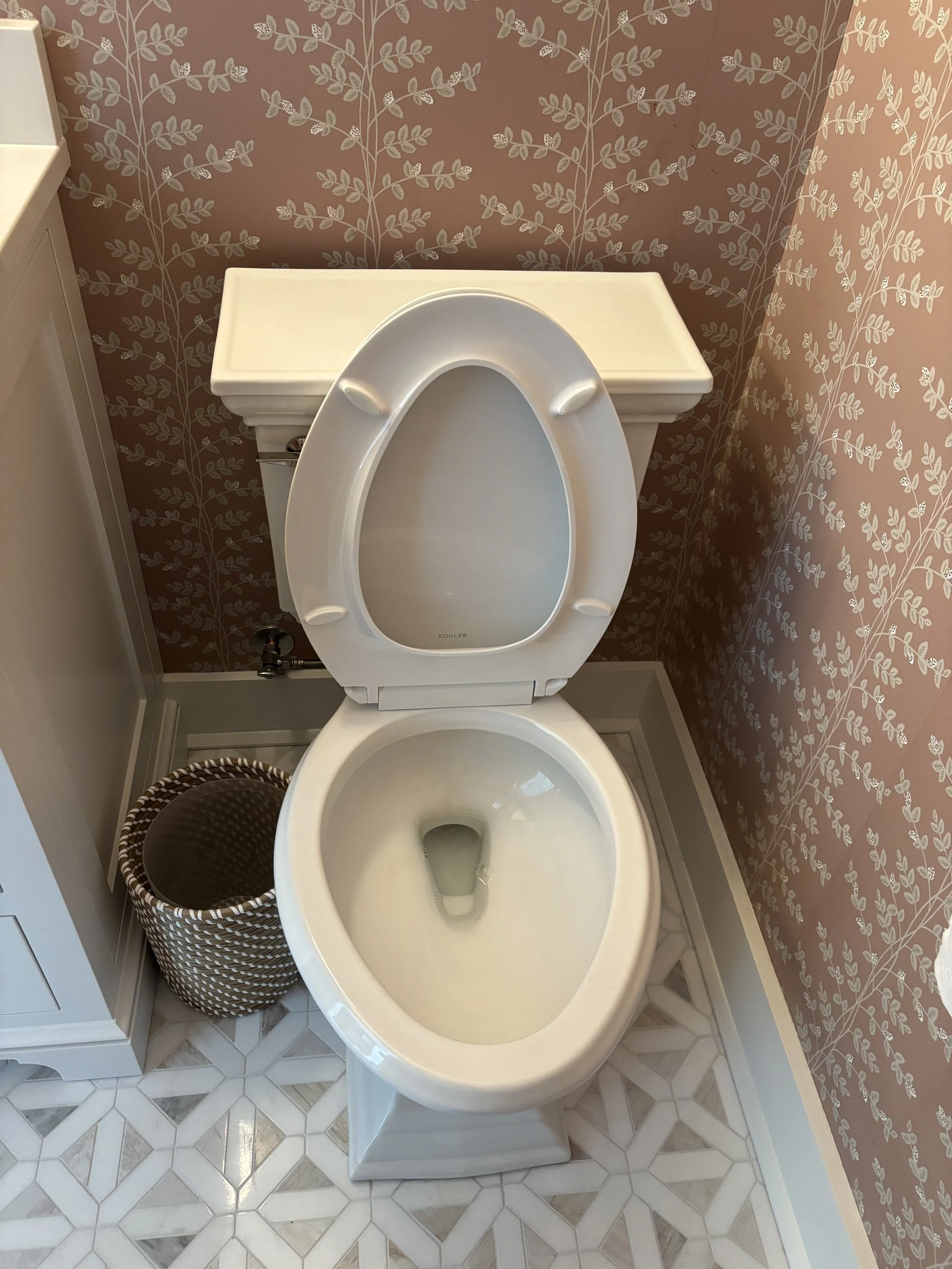 Empty white toilet with the lid open against a pink floral wallpapered bathroom wall, with a small basket underneath and a white cabinet to the side.