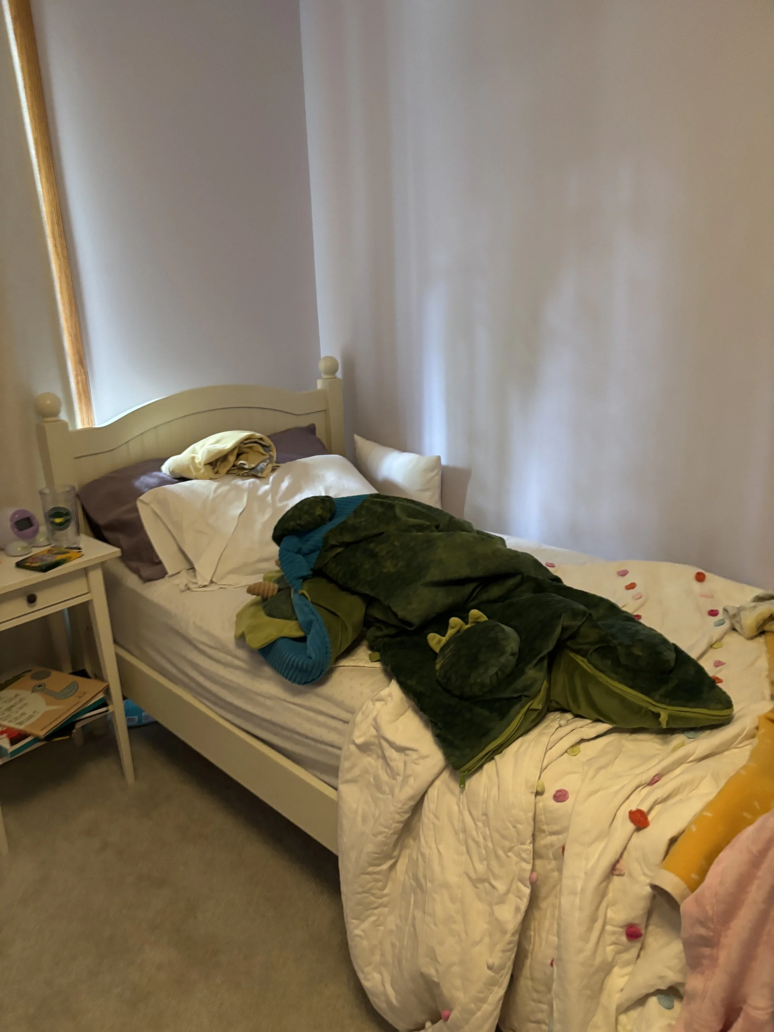 A bedroom with a white bed frame and a bed covered in a cream-colored quilt. There are clothes and blankets crumpled on the bed. Next to the bed is a small white nightstand with various items on it, including books, water bottles, and a clock. The wa