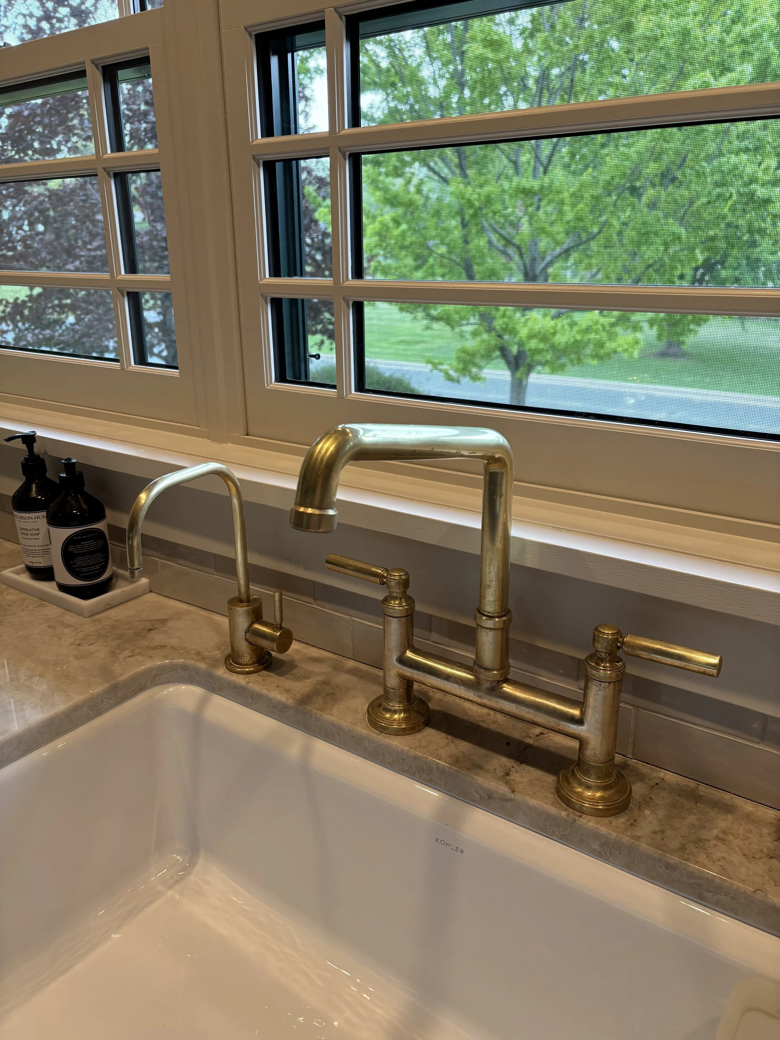 View of a kitchen sink with an exposed gold-colored faucet beneath a window showing green trees outside. Bottles of soap are near the sink.
