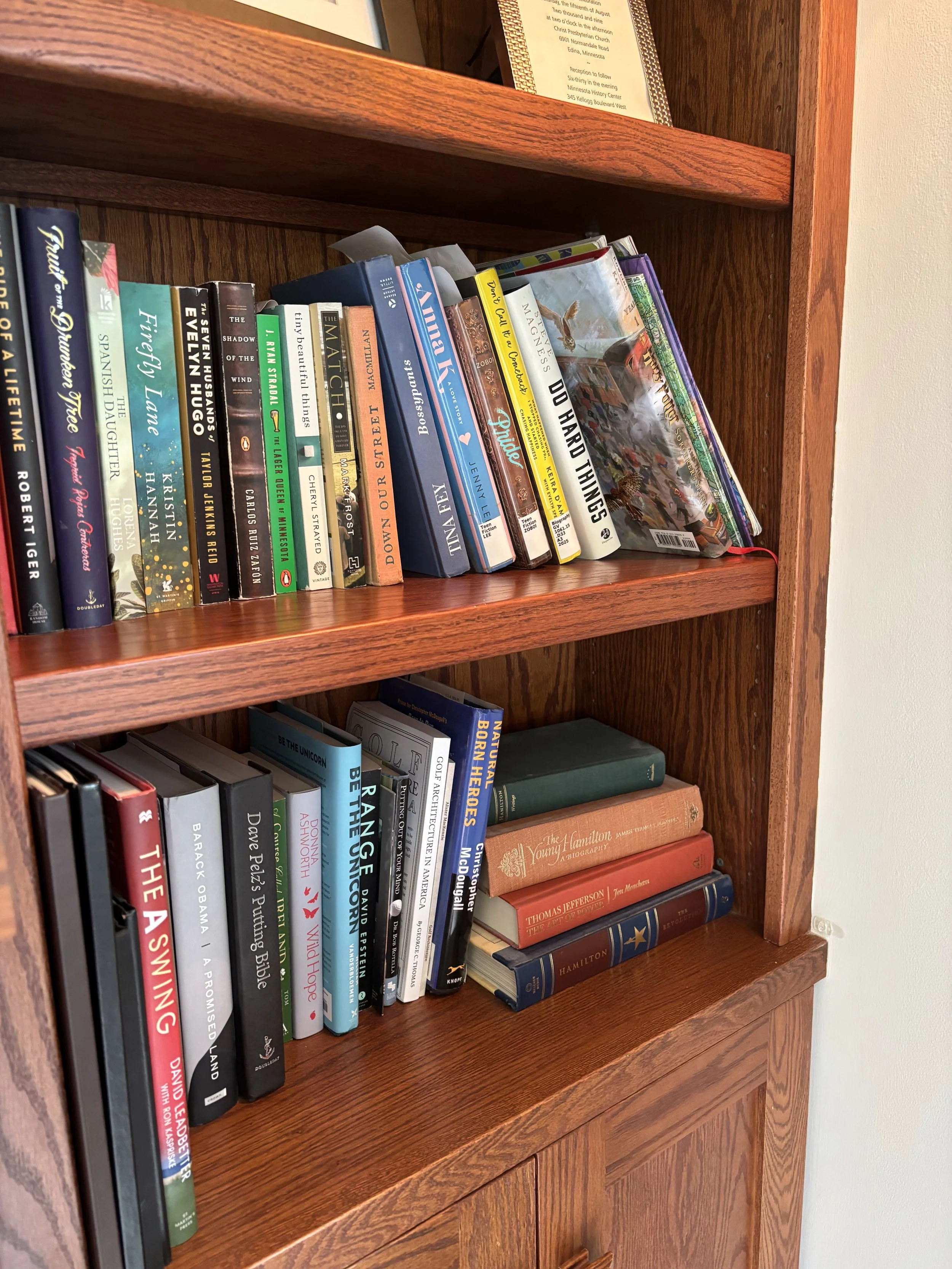Wooden bookshelf filled with various books, some standing upright and others stacked horizontally, with a framed document or certificate visible on the top shelf.