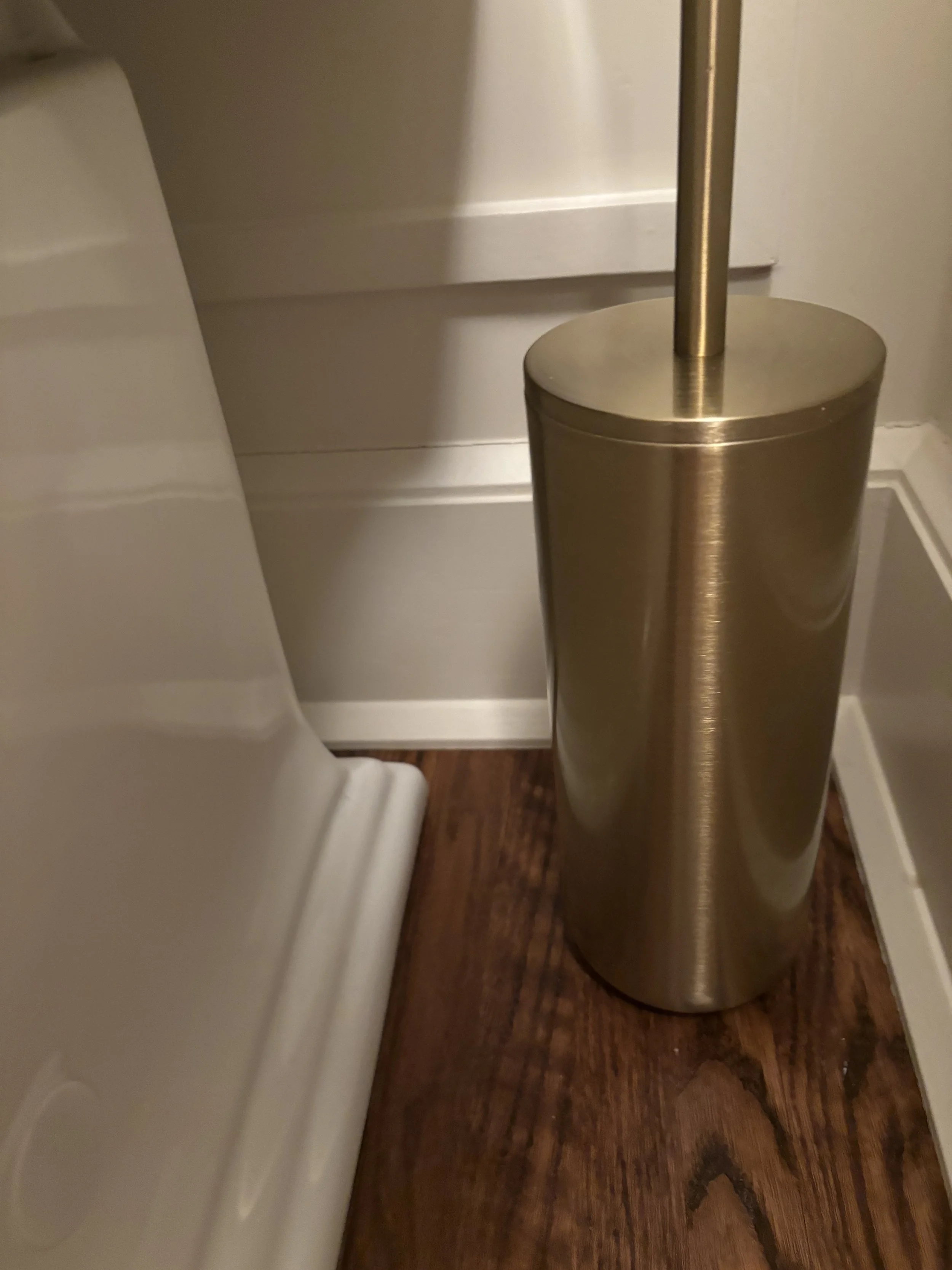 A close-up photo of a metallic toilet paper holder and part of a white toilet and white shelving unit on a hardwood floor.