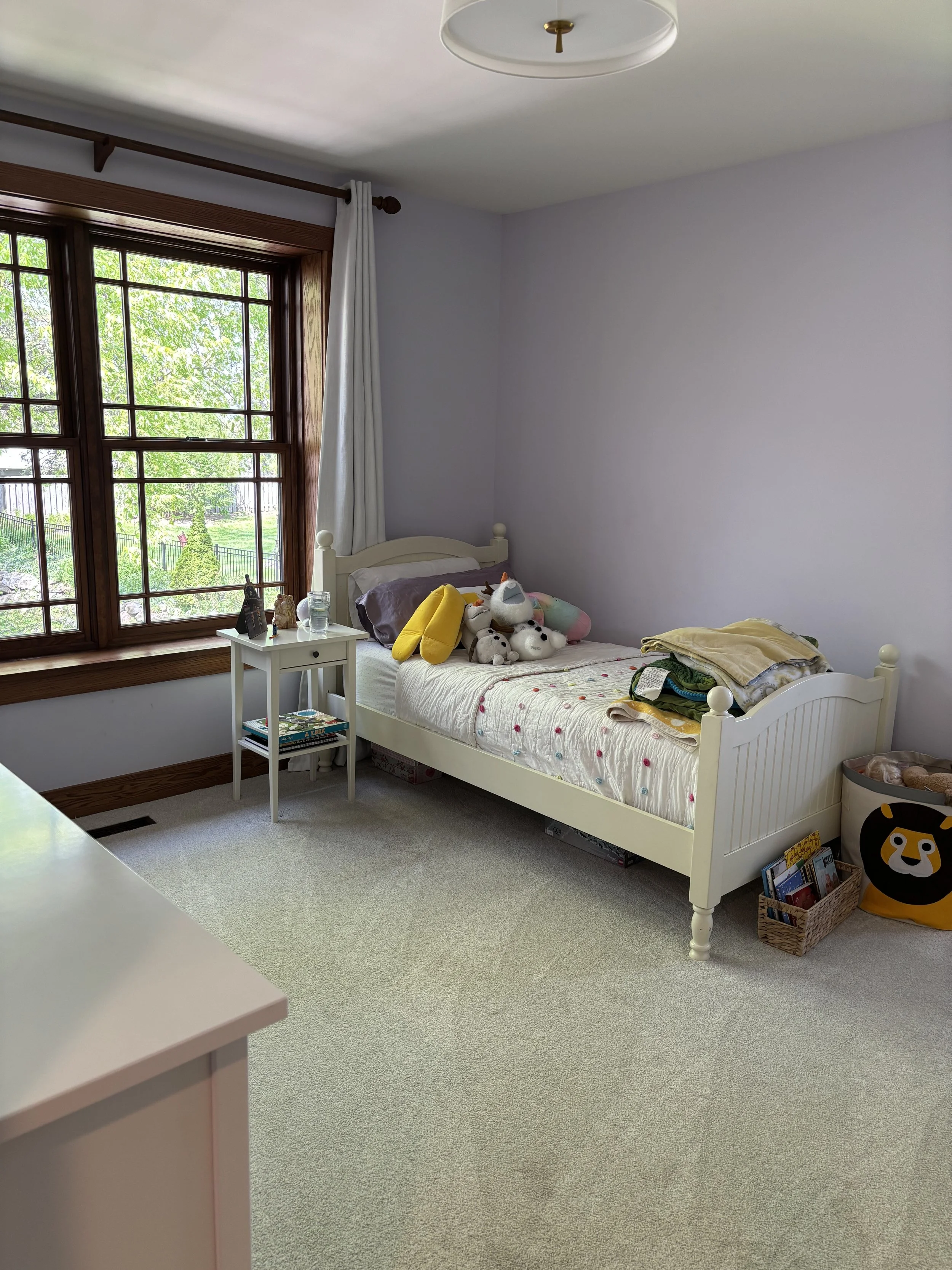 A child's bedroom with a single white bed, stuffed animals, books, and blankets, near a large window with white curtains, and a beige carpet.