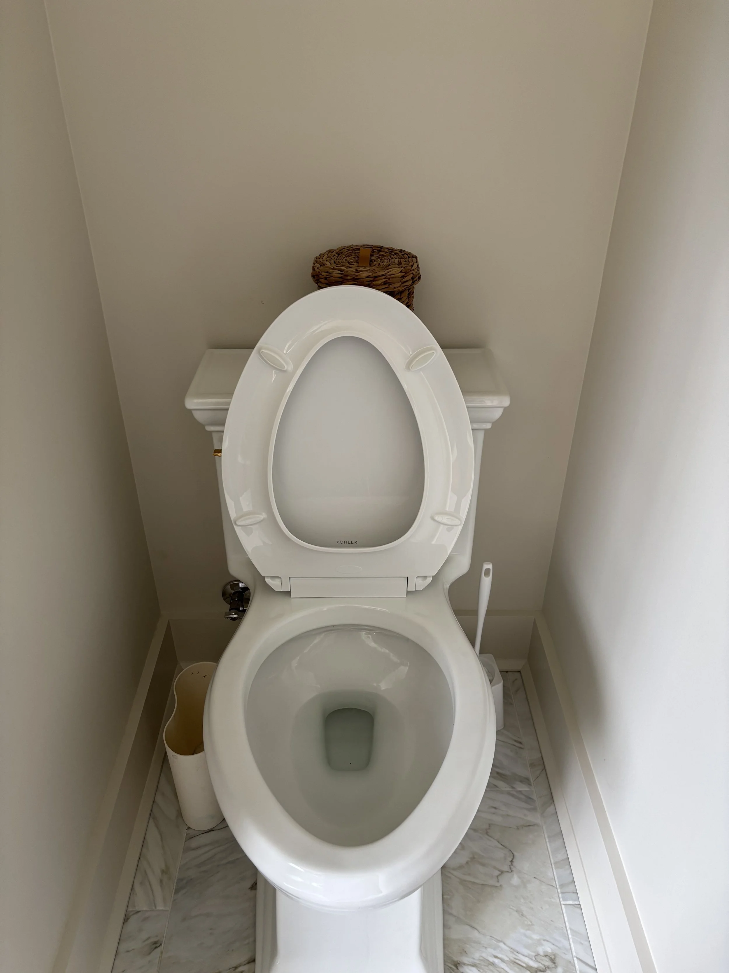 Empty white toilet with lid up in a small bathroom with beige walls and marble floor, a small trash bin to the left, and a woven basket behind the toilet.