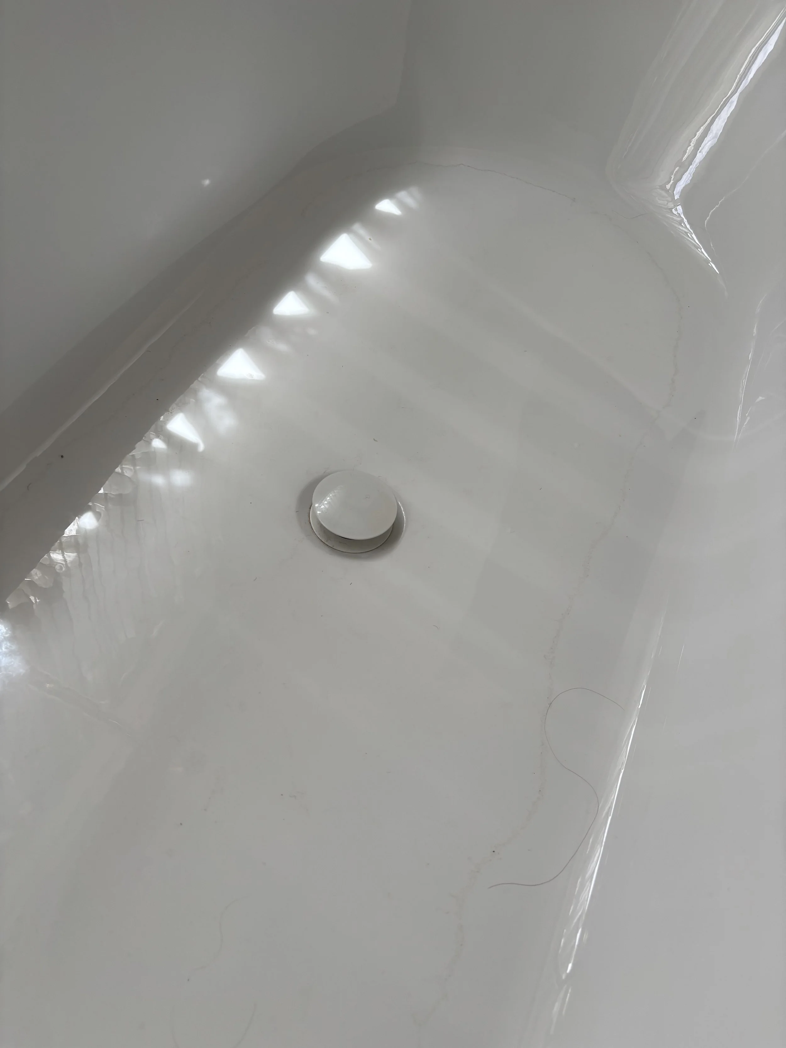 Close-up of a white bathtub with a drain and a shiny vertical pipe, with sunlight reflections creating patterns on the surface.
