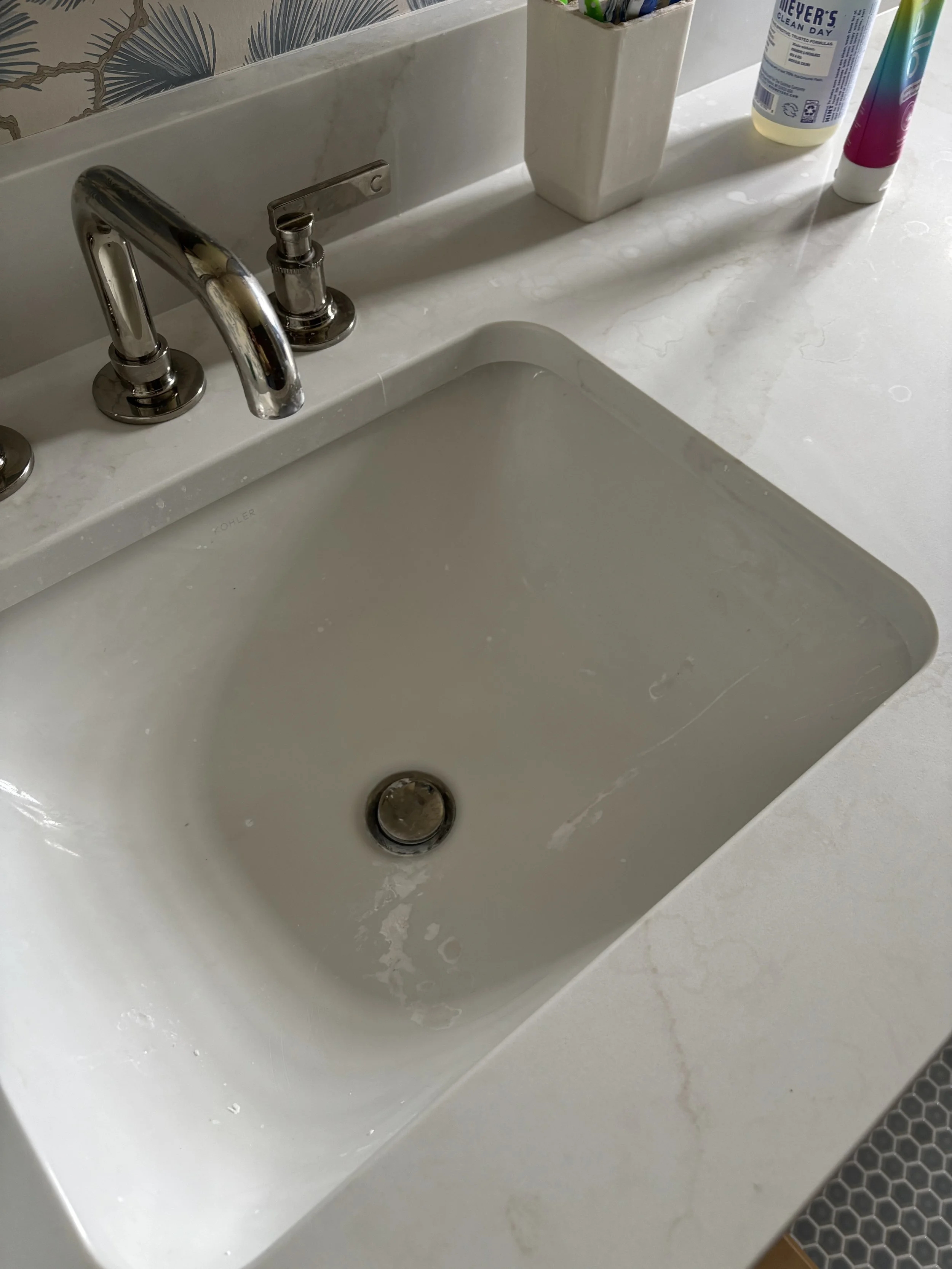 Bathroom sink with silver faucet, soap dispenser, toothbrush holder, and bottles of cleaning products on the counter.
