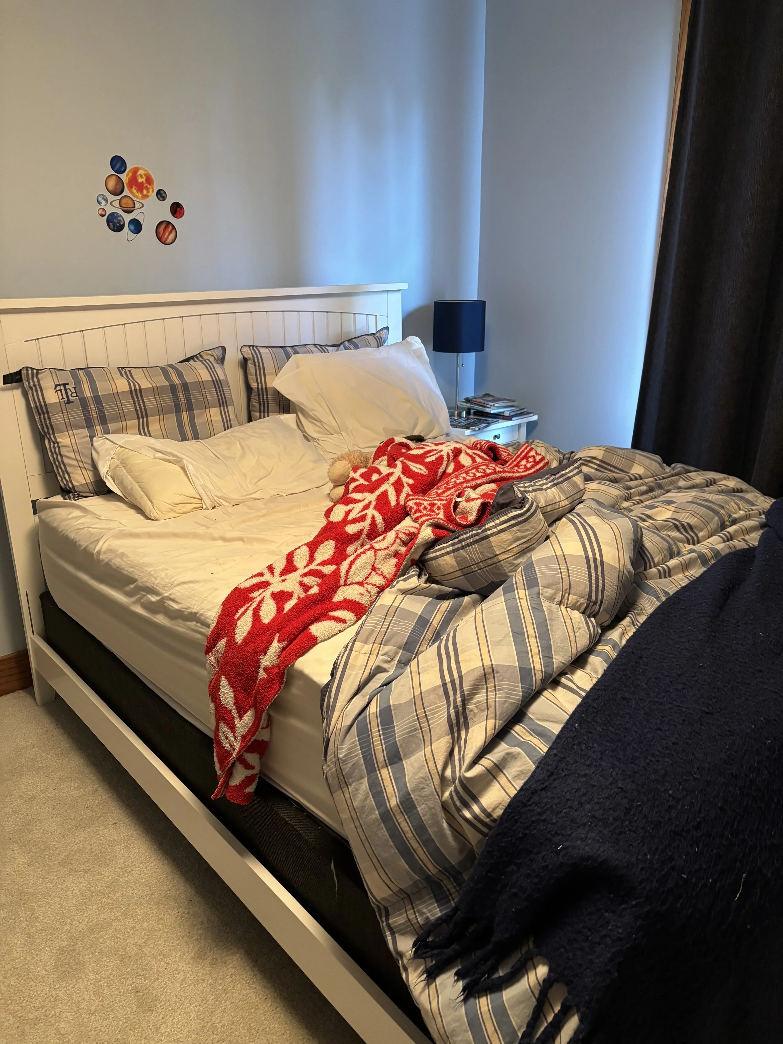 A messy bed with plaid sheets, multiple pillows, a red and white blanket, and a teddy bear, next to a small bedside table with magazines and a black lamp, in a bedroom with a light blue wall and planet stickers.