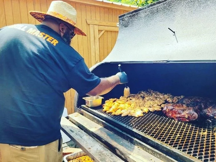 A person wearing a straw hat and a dark blue shirt is grilling chicken and ribs on an outdoor barbecue grill, with a wooden fence in the background.