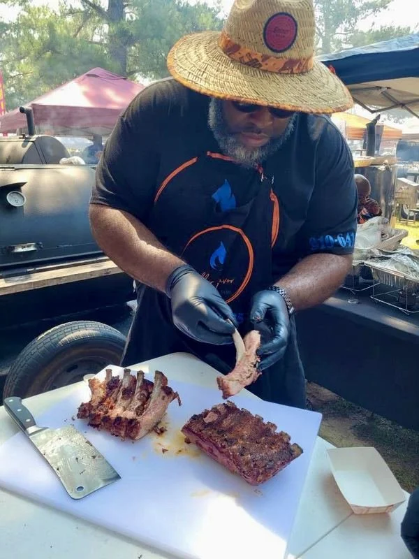 Man wearing straw hat, sunglasses, and black gloves preparing ribs at an outdoor barbecue event, with a grill and tents in the background.