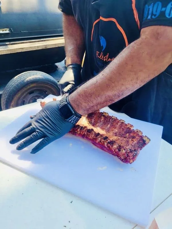 Person wearing black gloves preparing or slicing a rack of ribs on a white surface outdoors.