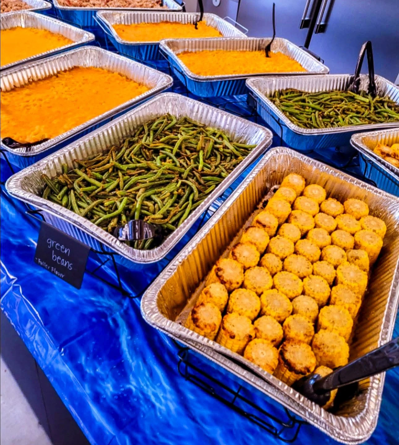 Food trays on a blue tablecloth buffet with green beans, baked macaroni and cheese, and breaded fried cauliflower.